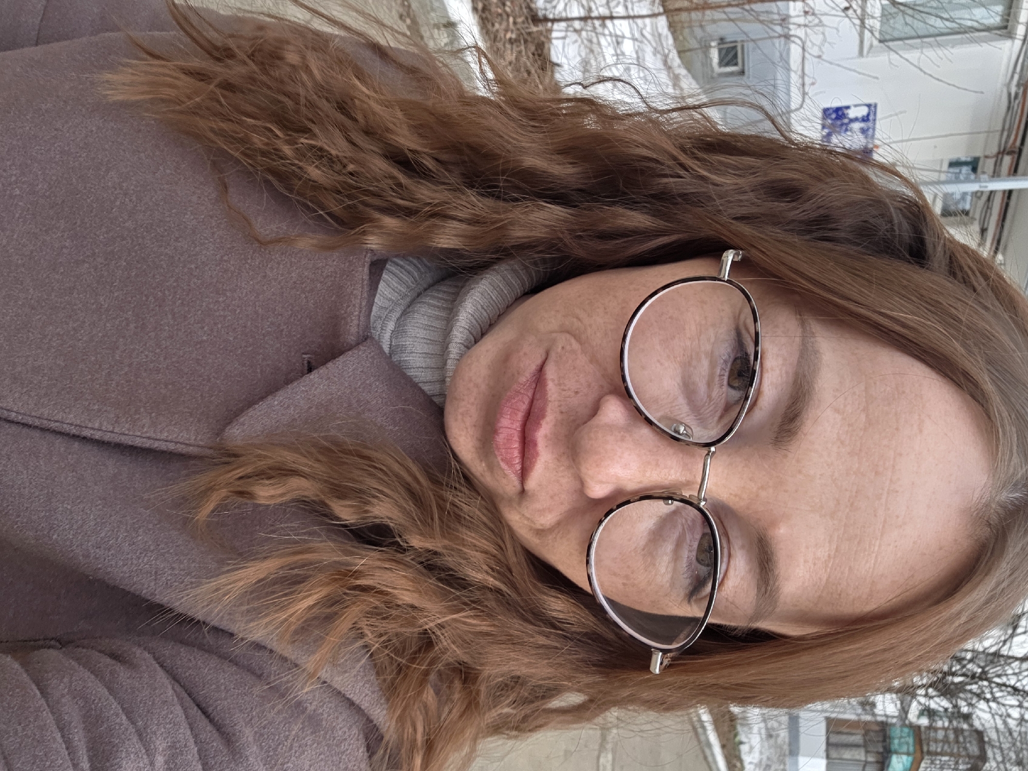 A fair-skinned female, approximately 30-45 years old, with long, wavy reddish-brown hair and visible freckles, directly faces the camera. She wears round eyeglasses with a dark and light patterned frame, a grey ribbed turtleneck, and a muted purple-brown wool coat. Her expression is serious. The scene is an outdoor urban environment in the city of Chaikovsky, Russia. In the background, a light-colored multi-story residential building is visible, featuring windows and exterior pipes or cables. A blue street sign with white Cyrillic lettering, partially obscured, is affixed to the building; the visible characters suggest it may read "ул. Мира" (Mira Street). Patches of melting snow cover the ground, alongside bare tree branches and sparse vegetation, indicating late winter or early spring conditions. A second building with green-paneled enclosed balconies is visible in the distant right background.