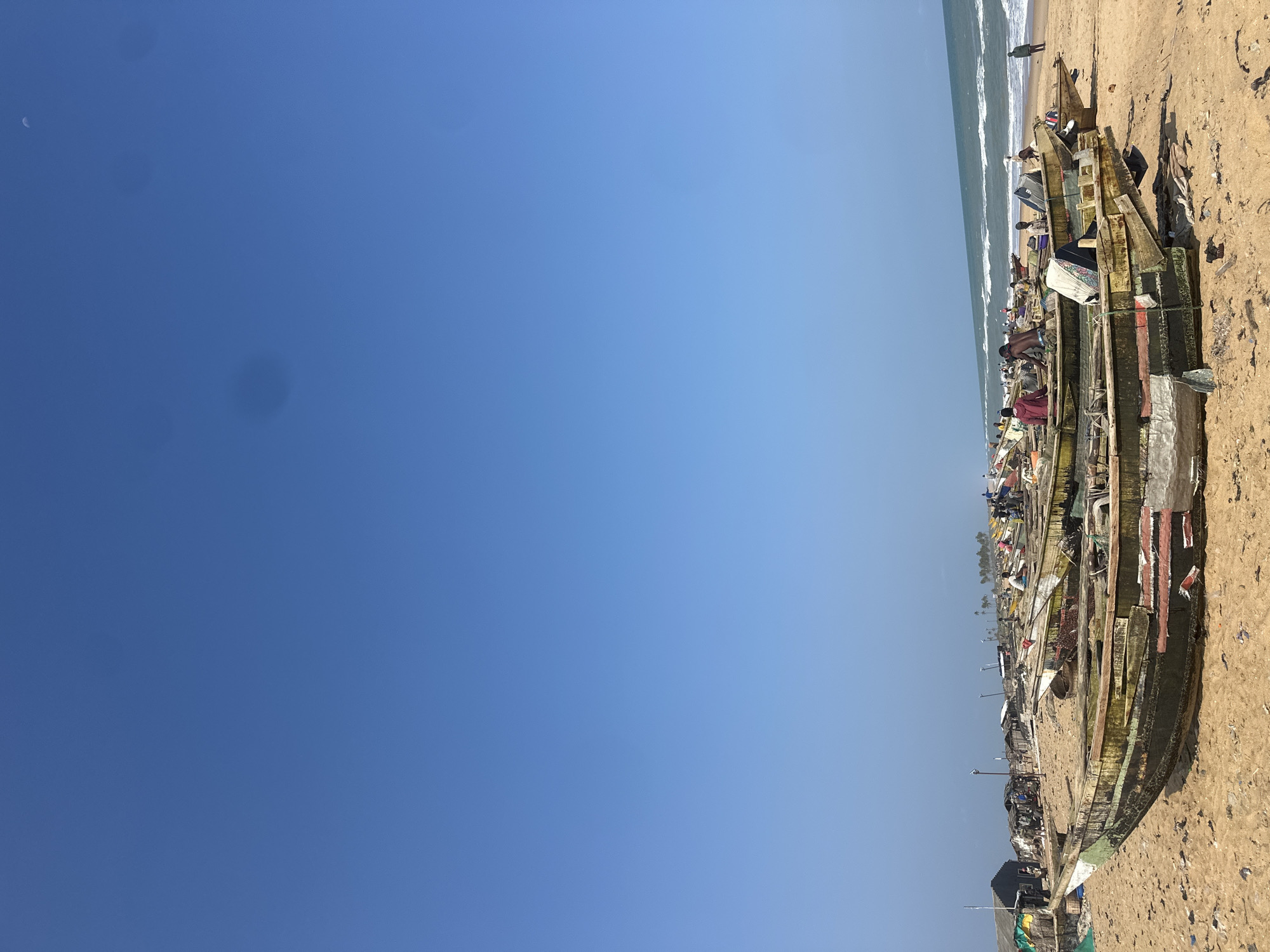 This is a photograph of a beach scene in Badagry, Nigeria. The image is oriented vertically, with the sky occupying the left two-thirds and the beach and ocean on the right. The sky is a clear, bright blue, with a faint crescent moon visible in the upper left corner. Scattered dark spots are present in the sky.
The beach is sandy and appears to be littered with debris. A line of several traditional wooden fishing boats is pulled up onto the sand. The boats are of varying sizes and appear weathered, with some showing signs of wear and tear, and painted with different colors. There are people visible in and around the boats. Some appear to be working on the boats or tending to them, while others are standing or sitting. The ocean is visible in the background, with small waves breaking on the shore. A single person is standing near the water's edge.
In the distance, along the coastline, there are structures that appear to be part of a settlement or village, with some buildings and possibly a row of palm trees. The lighting suggests it is daytime, likely morning or afternoon given the clear sky. The overall impression is a working fishing community on a beach. No visible text is present in the image.