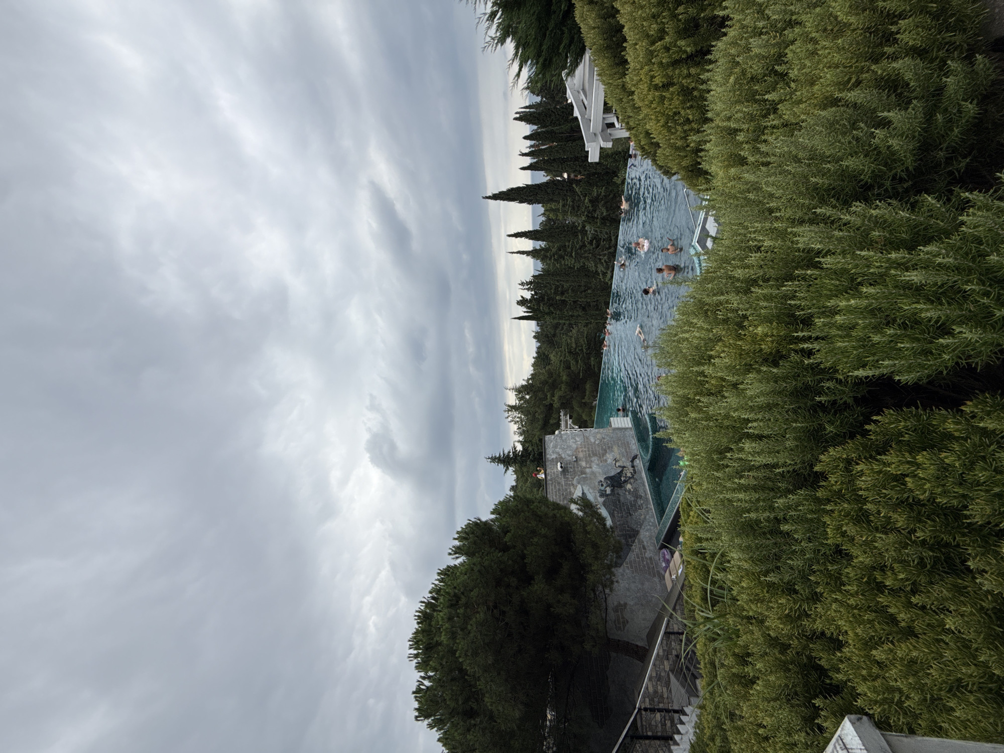 The image shows an outdoor infinity pool nestled among lush green trees and bushes, with a cloudy sky overhead. The pool is filled with clear blue water, and several people are swimming and relaxing in it. The pool is situated on a hillside, offering a panoramic view of the surrounding landscape, which includes the sea in the distance. The architecture surrounding the pool is modern and sleek, with clean lines and minimalist design. There is also a large, mature pine tree to the left of the pool, adding a natural element to the scene. The overall impression is one of a tranquil and luxurious retreat, a perfect place to escape the hustle and bustle of city life. The location is identified as Yalta, Ukraine.