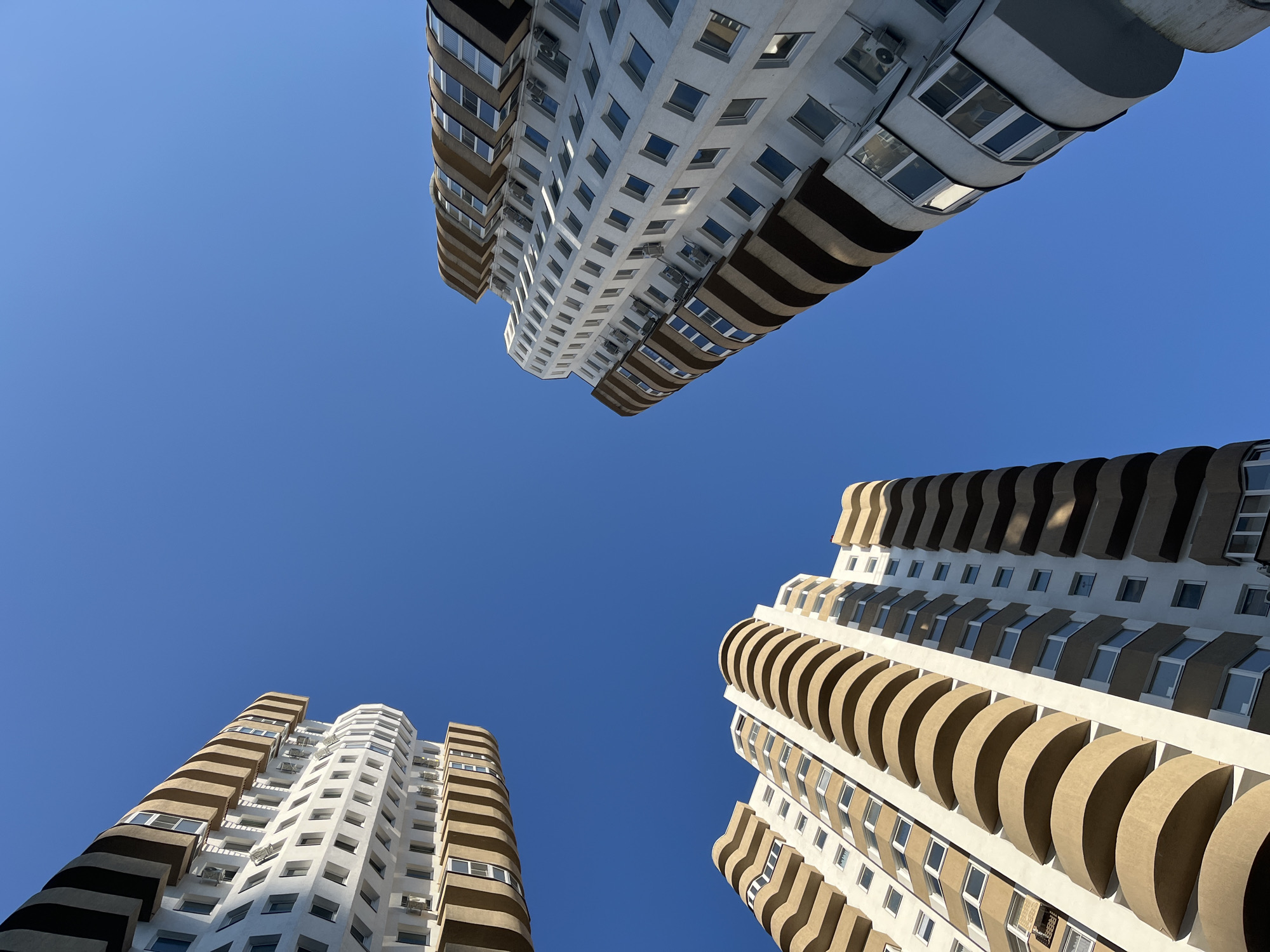 Three tall residential buildings with modern architecture are captured from a low angle, looking up towards a clear blue sky. The buildings feature white facades accented with curved, beige balconies that create a distinctive, wavy pattern. Some windows are visible, as are air conditioning units mounted on the exterior walls. The buildings appear to be located in Sochi, Russia, a Black Sea resort city known for its diverse architecture. The scene is devoid of people or any visible activity.