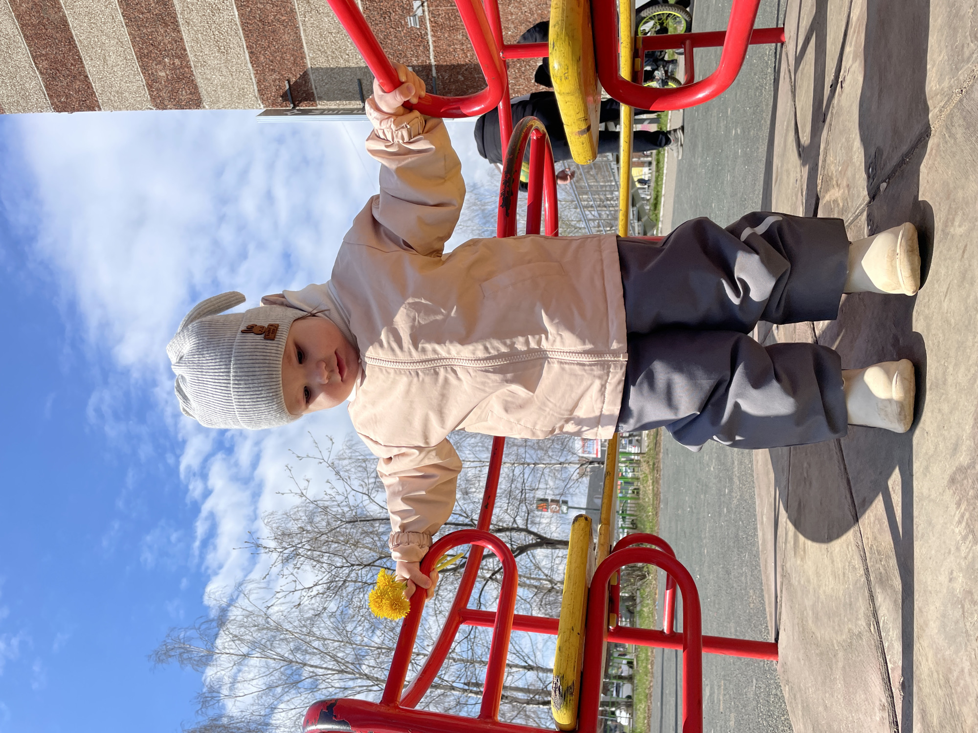 A young child stands on a vibrant red and yellow playground structure under a clear, bright blue sky with scattered white clouds. The child is dressed in a light pink or beige jacket, gray pants, and a light gray knit hat adorned with bunny ears, completed with light-colored boots. They are holding onto a red bar of the equipment with both hands, and their right hand also holds a small bunch of yellow dandelions. The child looks towards the camera with a calm expression.
The outdoor setting features bare trees in the background, suggesting early spring. The ground is a paved or asphalt surface, with distinct shadows indicating bright daylight. In the upper left portion of the image, a building with horizontal brown and beige stripes is visible. Faintly in the deeper background, other elements of the playground, including what appears to be part of a bicycle wheel and another person's legs, can be discerned. The scene depicts a sunny day at a playground in Izhevsk, Russia. No discernible text is visible.
