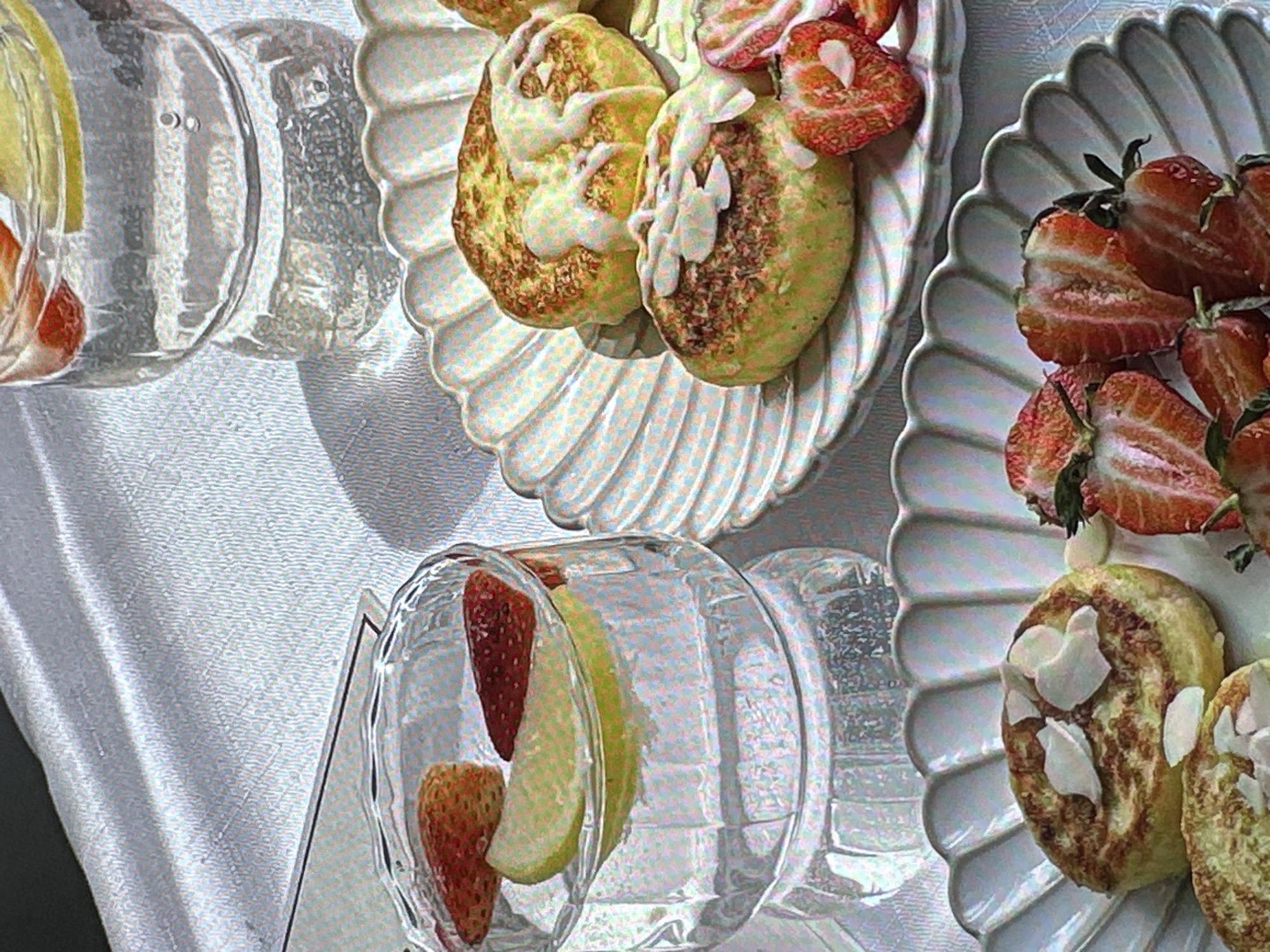 The image shows a close-up of food and drinks on a white tablecloth. There are two plates of what appear to be pancakes or crumpets, topped with different sauces and fresh, cut strawberries. Alongside the plates are two glasses filled with a clear liquid, slices of lemon, and whole strawberries. The scene likely represents a breakfast or brunch setting. The location and association with the city of Yerevan, Armenia, cannot be determined from the visual information.