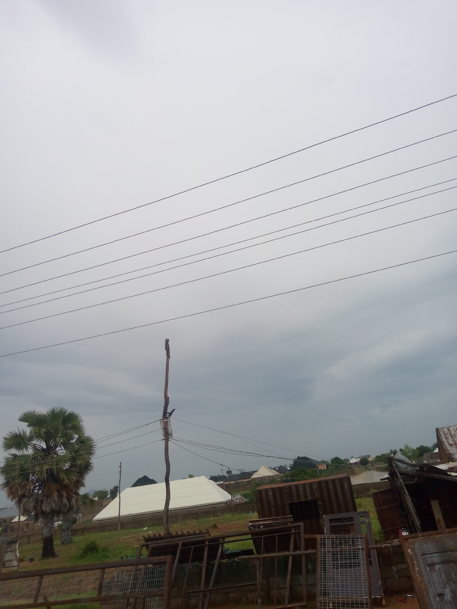 The image depicts an outdoor scene in Kona Garu, Nigeria, under a cloudy sky. In the foreground, a rusty metal fence is visible, with various metal structures and debris behind it, including corrugated iron sheets and what appears to be discarded doors or gates. Further back, a palm tree stands prominently on the left side of the frame, with dry fronds. To the right of the palm tree, a white-roofed building with a dark, pointed structure on its peak is situated. In the mid-ground, several other buildings with similarly colored roofs and a metallic shipping container are visible, surrounded by green vegetation. Overhead, multiple power lines stretch across the frame, supported by a wooden pole with some attached wires and hardware. The overall environment suggests a semi-urban or rural setting with a mix of residential structures and possibly some form of informal industrial or storage area.