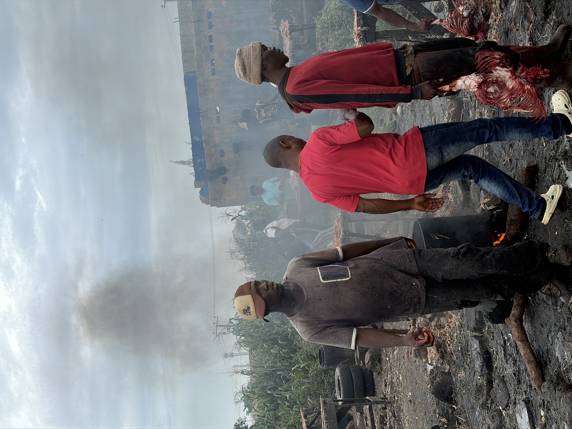 This image depicts an outdoor scene in a location that appears to be a processing or market area, potentially in Bukuru, Nigeria, given the provided context. The environment is somewhat unkempt, with muddy ground, debris, and vegetation, including what looks like corn stalks, visible. Smoke billows in the background, originating from an unseen source and obscuring parts of the sky and a structure resembling a building or large container.
In the foreground, several individuals are present. On the left, a man wearing a grey t-shirt and a baseball cap stands looking off to the side. He appears to have a reddish stain on his hand. To his right, another man in a bright red t-shirt and jeans is walking away from the camera. Further to the right, a man in a red hooded sweatshirt and a beanie is walking, carrying what appear to be raw, plucked chickens. Another person in a blue shirt and jeans is partially visible on the far right. In the background, other figures are seen near the smoky structure. There are also objects like tires and barrels present in the scene, and a fire is visible in a barrel. The overall impression is one of a busy, somewhat chaotic, and possibly industrial or agricultural activity.