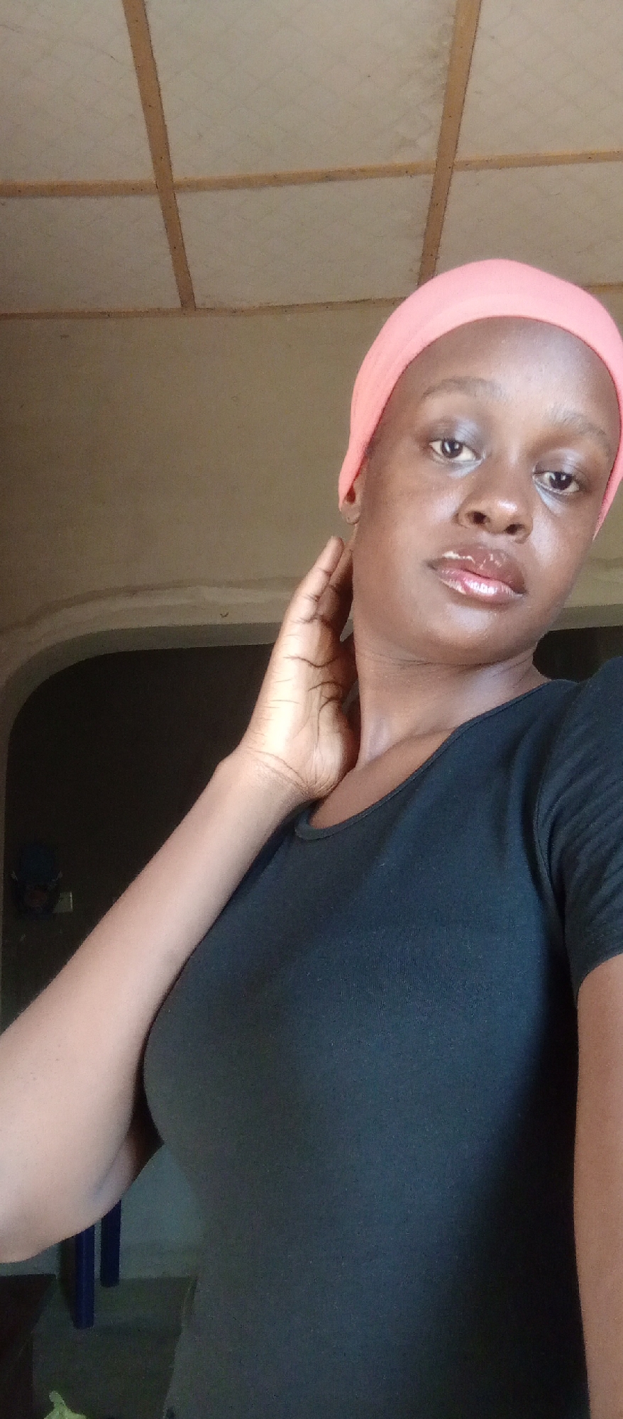 A medium shot depicts a dark-skinned young woman indoors. She is wearing a pink headscarf and a black top. Her right hand is raised near her face, with her fingers lightly touching her neck. A dark, arched doorway is visible in the background. The lighting is natural, casting shadows and highlights on her face and clothing. The location is Akwanga, Nigeria.
