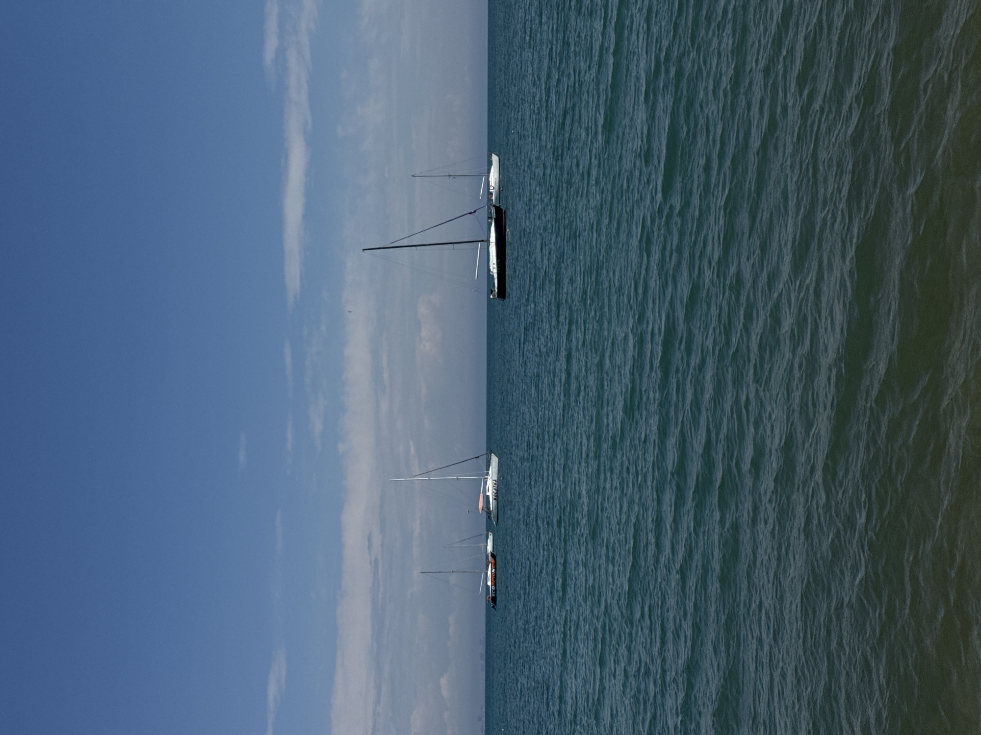 The image shows several sailboats on the Black Sea, with a clear blue sky and some wispy clouds visible. The location is likely near Yevpatoriya, Ukraine, based on the prompt. The water has small waves. There are three sailboats visible, and they seem to be relatively still.