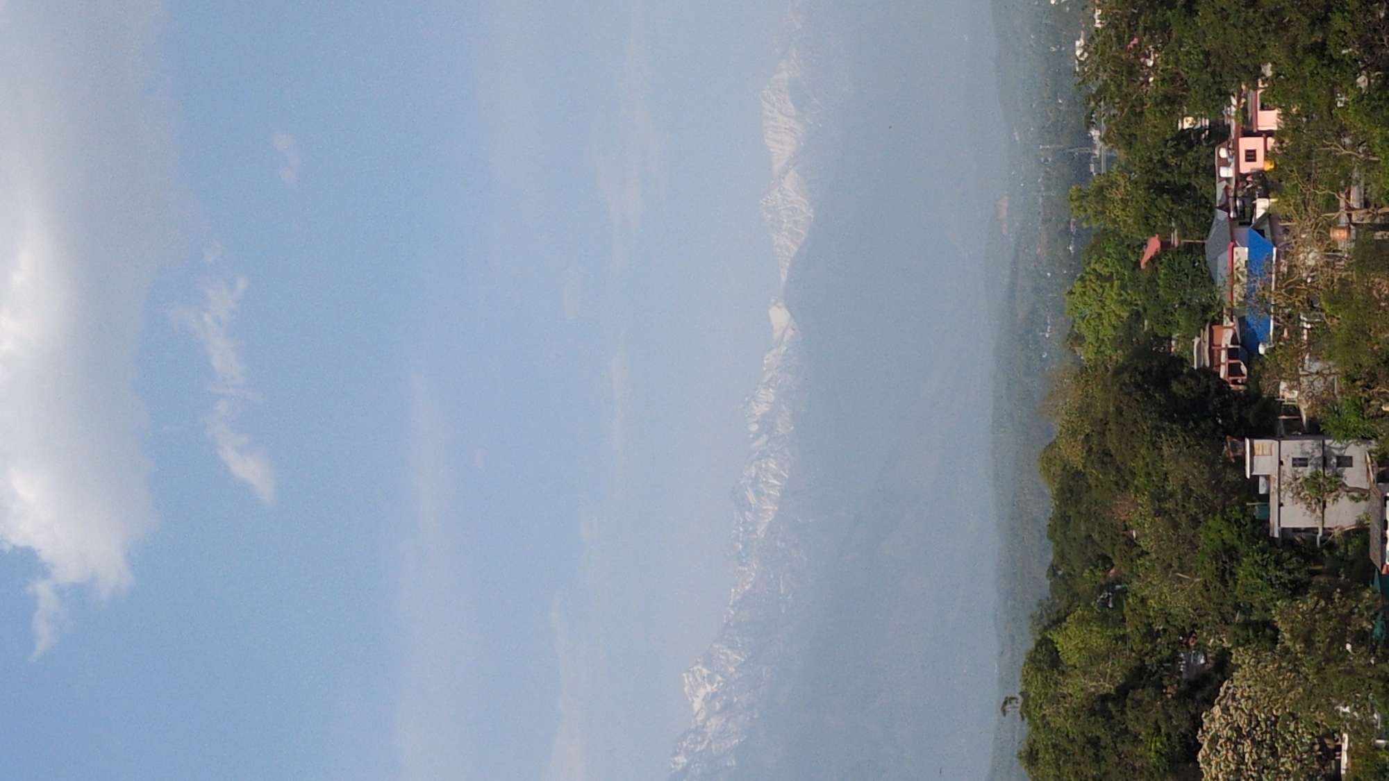 The image shows a view of a mountainous region in Kangra, India. The left side of the frame is dominated by a light blue sky with scattered clouds. A mountain range is visible in the center, its peaks shrouded in a hazy mist. The right side of the image depicts a densely vegetated area with trees interspersed with buildings. Several buildings can be seen amongst the trees, varying in color, with a few notable buildings having pink, blue, and white exteriors.