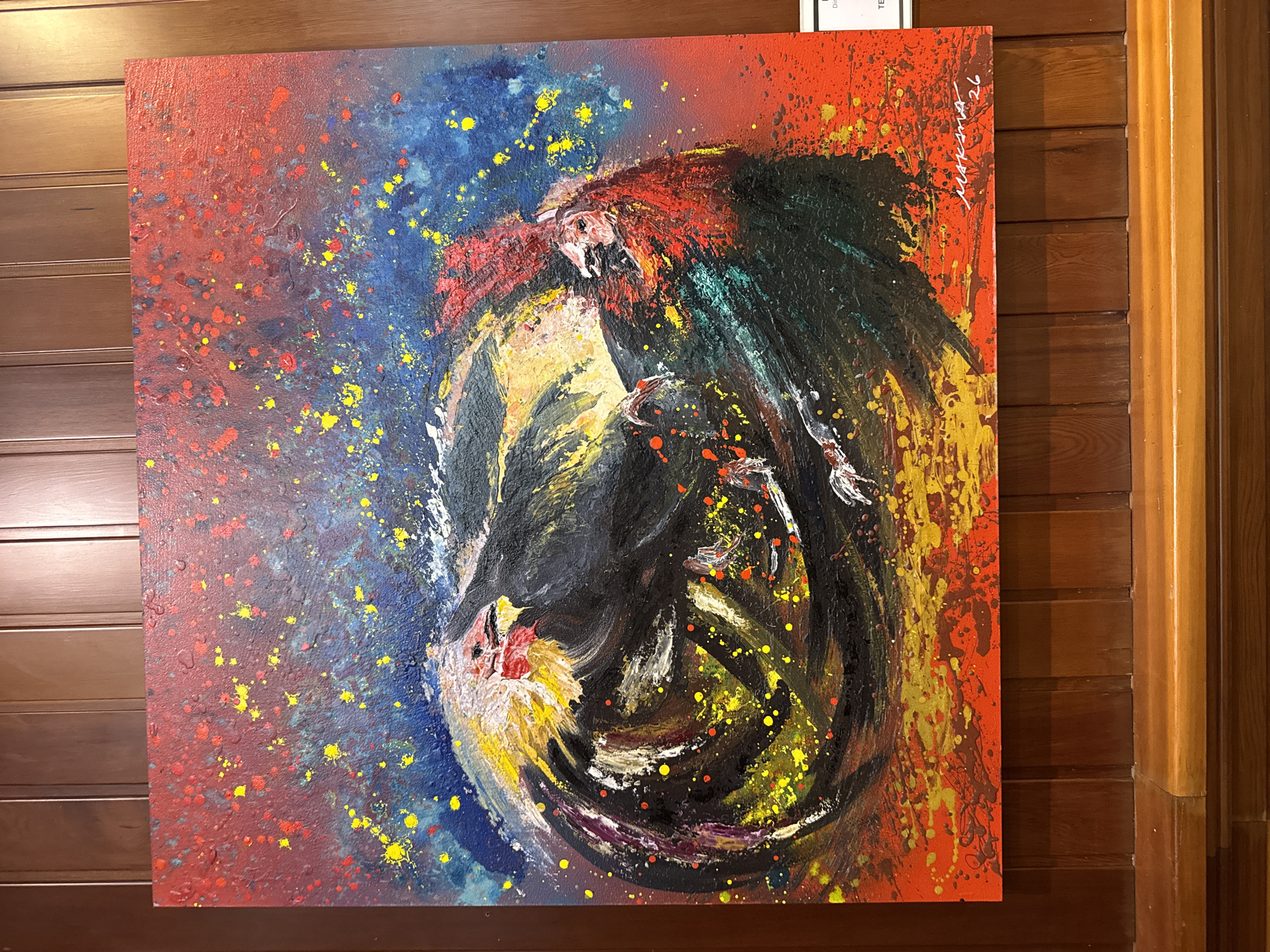 This is a photograph of an abstract painting featuring two roosters in a circular motion against a vibrant, speckled background of red, blue, and yellow. The painting is hung on a dark wooden paneled wall. The roosters are depicted with energetic brushstrokes, with dominant black and red colors, and splashes of yellow and white on their heads and tails. The background is textured with a splatter effect of yellow and red dots on a gradient of blue to red. The signature "MAKM 26" is visible in white on the lower right corner of the painting. A small white card is partially visible on the right edge of the frame, suggesting the painting is on display in a gallery or exhibition space, potentially in Torrejón de Ardoz, Spain. The painting is illuminated by artificial light, casting subtle shadows.