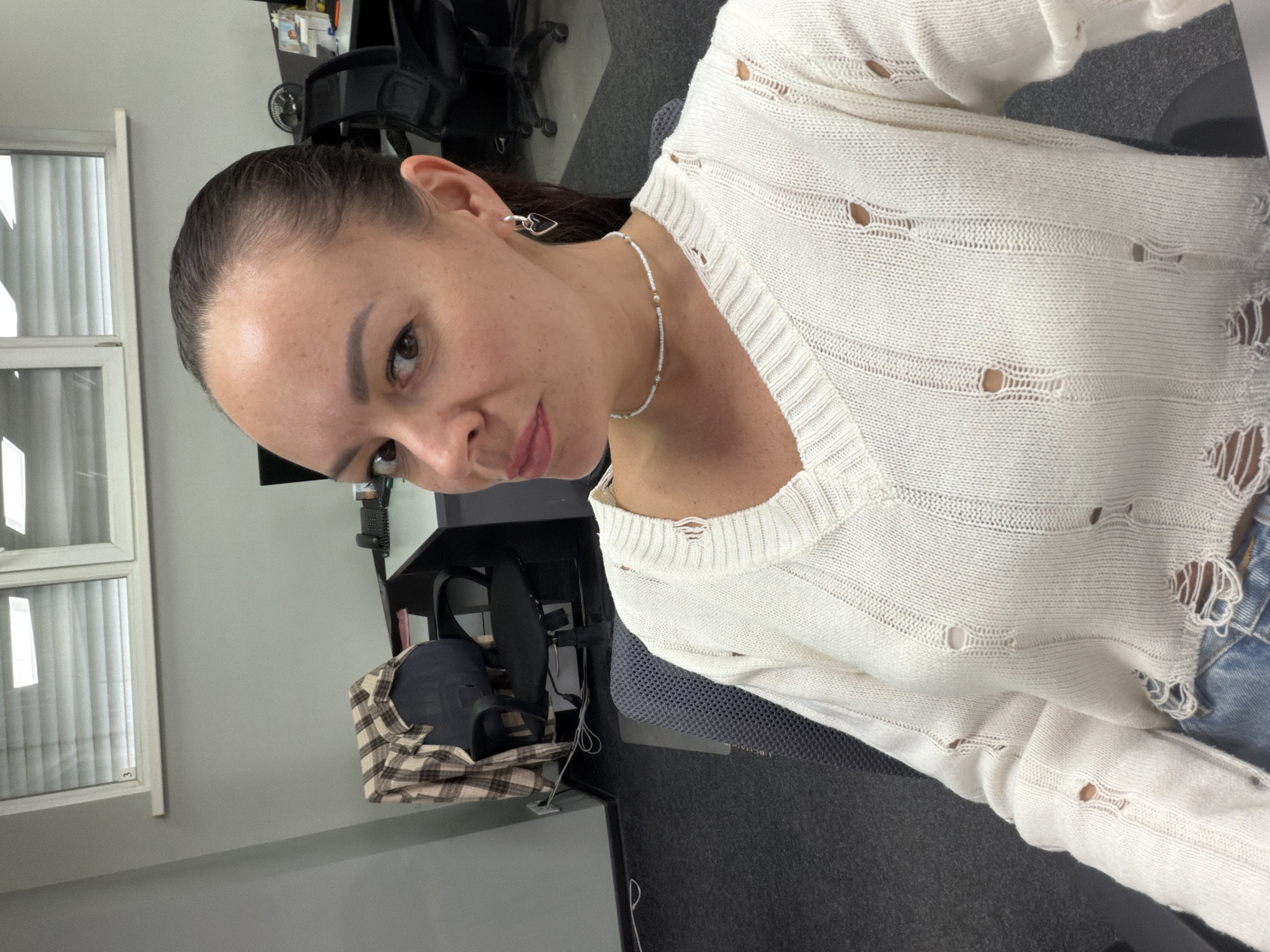 A woman with brown hair pulled back in a ponytail is facing the camera with a slight tilt. She is wearing a white, distressed v-neck sweater and a beaded necklace. Her earrings are visible. The background shows an office setting with a window to the left, office equipment including chairs, desks, and a monitor. The location is indeterminate.