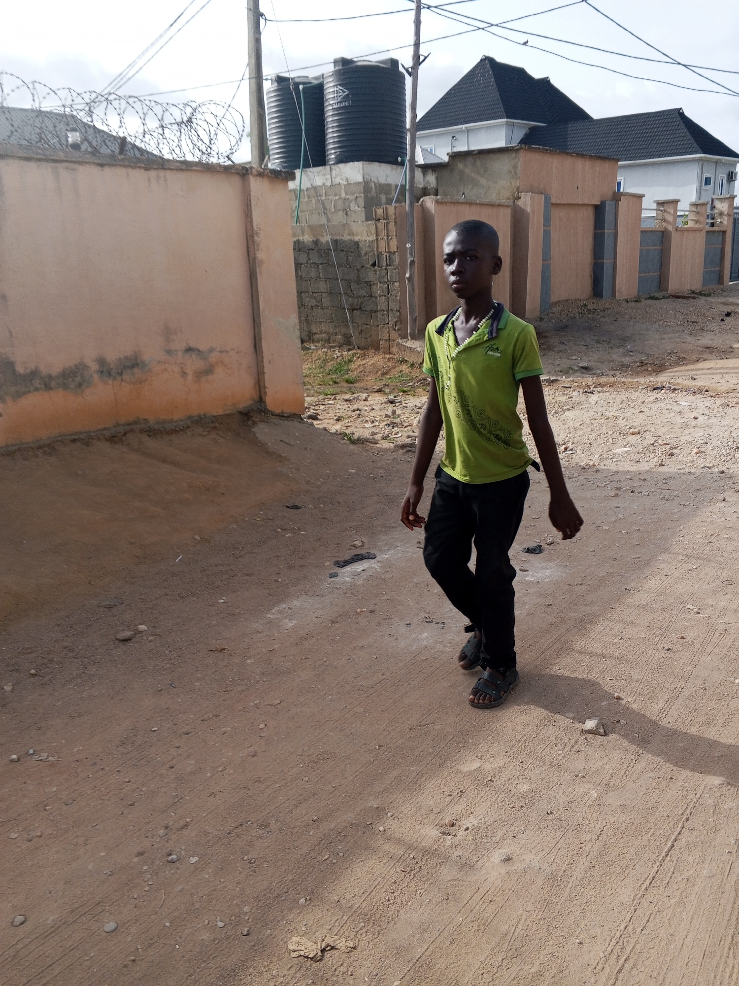 A young boy walks on a dirt road in Otokiti, Nigeria. The road is dry and dusty, with vehicle tire tracks visible. The boy wears a green t-shirt, black trousers, and dark sandals. In the background, there's a walled compound with barbed wire on top, water tanks, and houses with dark roofs. The scene suggests a residential area.