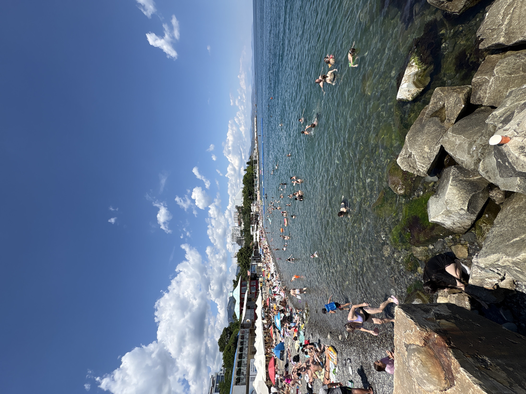 This image depicts a crowded beach on a sunny day in Alushta, Ukraine. The scene is dominated by a large expanse of the sea, with a pebble beach packed with sunbathers and swimmers. The water is clear and shallow near the shore, gradually deepening further out. Numerous people are engaged in various activities: swimming, sunbathing on towels and loungers, and wading in the water. Some individuals are seen with inflatable toys.
Along the beach, there are rows of white umbrellas providing shade, and several buildings, likely cafes or shops, are visible in the background. The landscape behind the buildings features green hills and some structures, suggesting an urban or resort area. The sky is a vibrant blue with scattered white clouds. Large rocks are present in the foreground, with some moss or algae visible on them, indicating their proximity to the water. A small red and white object, possibly a marker or a piece of debris, rests on the rocks in the bottom right corner. The overall atmosphere is one of a busy summer day at a popular tourist destination.