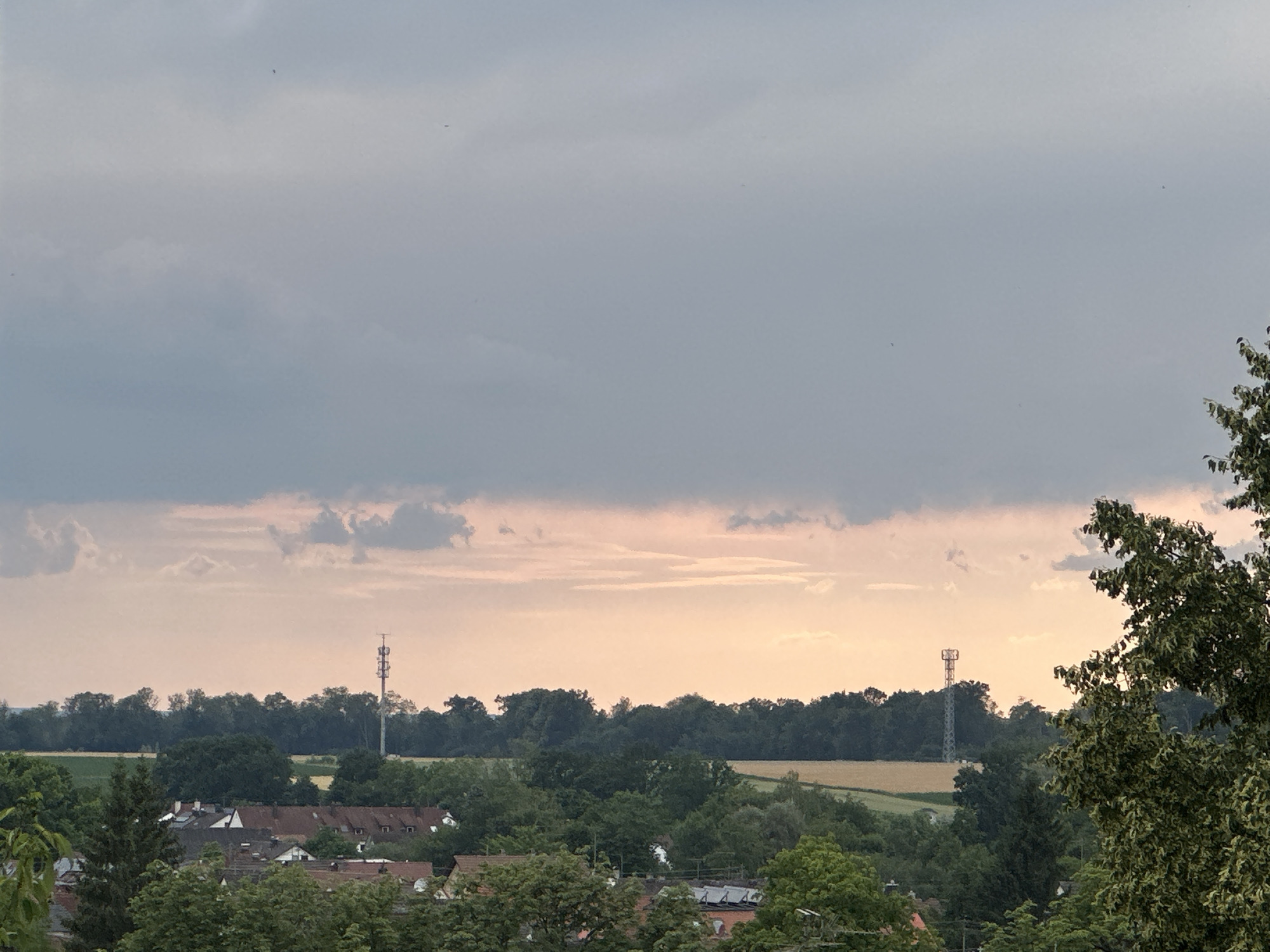 The image depicts a rural landscape with a village nestled amongst trees under a cloudy sky. In the mid-ground, there is a cluster of buildings with red-tiled roofs. Two tall, lattice-style communication towers are visible, one more centrally located and the other to the right. Fields of crops, possibly wheat, are interspersed with dense green trees that form a line across the horizon. The sky is filled with a mix of dark gray and lighter, pinkish-orange clouds, suggesting either sunrise or sunset. The foreground features lush green trees, partially obscuring the view of the village. There are no people or animals visible in the scene. The overall impression is one of a tranquil, natural setting with man-made structures present.