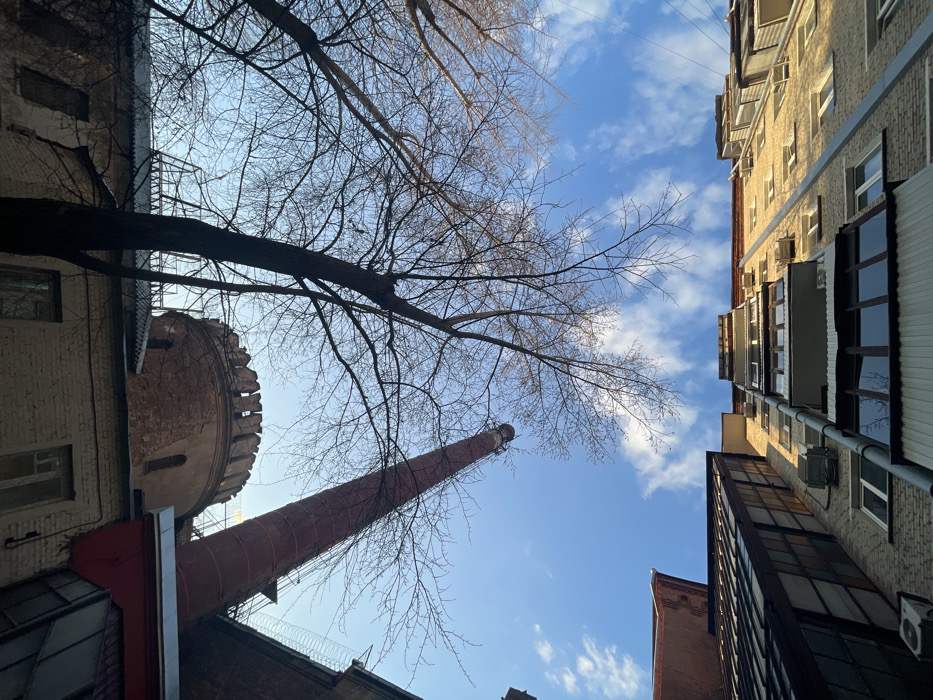 A low-angle, wide-perspective image captures a view upwards from a confined urban space in the Red October (Krasny Oktyabr) complex in the Zamoskvorechye district of Moscow, Russia. The sky, a vibrant blue with scattered white cumulus clouds, dominates the central frame.
Framing the sky are several buildings. On the left and top edge, a multi-story building constructed of light-colored brick features numerous windows and several small balconies. A drainpipe runs vertically down this facade. Directly opposite, on the right and top edge, another multi-story building, appearing darker in tone, also presents multiple windows.
In the lower and mid-ground, a distinctive architectural ensemble is prominent. A round, beige-colored brick tower with crenellated battlements at its summit is visible. To its right, a tall, slender, red brick industrial chimney stack rises steeply, with an external ladder or scaffolding system affixed to its side. A section of concertina wire is visible on a rooftop or wall adjacent to the chimney. Below these structures, portions of other lower-rise buildings with windows and pitched roofs are visible.
Several bare deciduous trees, devoid of leaves, stand between the buildings, their dark, intricate branches silhouetted against the bright sky. No people, animals, or active interactions are present in the scene. The overall environment suggests a former industrial or historical site repurposed for other uses.