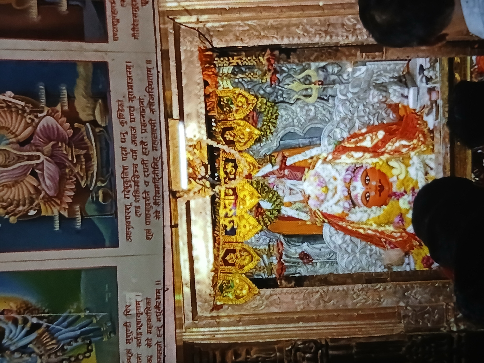 The image shows a Hindu shrine, possibly located within a temple in Bengaluru, India. The shrine features a decorated statue of a deity, possibly Hanuman or another powerful god, adorned with flowers, garlands, and colorful clothing. The statue is placed against an ornate, carved backdrop that includes trident symbols. The shrine itself is enclosed within a decorative frame, likely made of stone or a similar material. To the left of the shrine are painted panels depicting other Hindu deities and religious texts. People are visible, including the back of a person's head near the right edge of the frame, suggesting that the shrine is in a public or accessible location where people are visiting. The overall environment appears to be dimly lit, with a focus on illuminating the shrine.