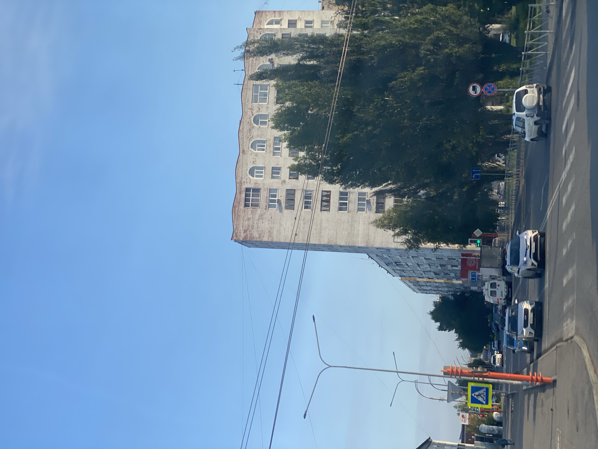 The image shows a street scene in Kemerovo, Russia, under a clear blue sky. A multi-story residential building with arched windows at the top dominates the background. In the foreground, a street with multiple lanes of traffic is visible, with several cars and a white ambulance driving on it. A pedestrian crossing sign is present on the left side of the road, and streetlights are installed along the sidewalk. Tall trees line the street, partially obscuring the view of the building. The overall impression is of a typical urban environment in Russia.