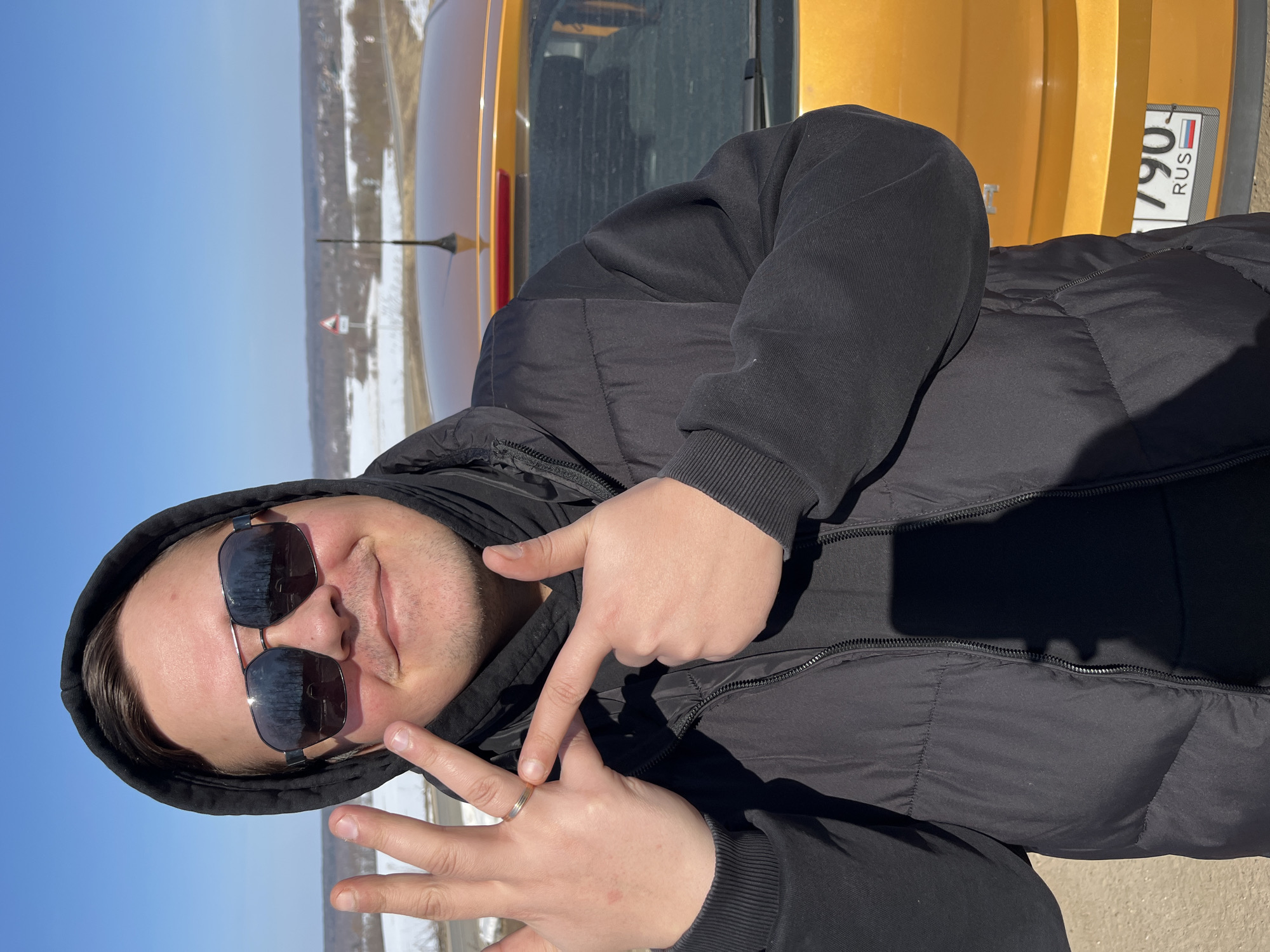 The image captures a male individual standing outdoors on a bright, sunny day in what is identified as Zubovo, Russia. The man, framed from mid-torso up, wears a black hooded jacket with the hood up, a black puffer vest, and dark, reflective aviator-style sunglasses. He presents with a slight mustache and stubble and is smiling towards the camera. His left hand is raised, with four fingers extended, prominently displaying a gold-colored ring on the ring finger. His right hand points with an extended index finger towards his raised left hand. Behind the subject, the rear section of a bright orange or yellow hatchback car is visible, featuring a roof antenna and a Russian license plate partially reading "790 RUS". The background reveals a clear blue sky above a broad, gently undulating landscape covered in patches of snow and dry vegetation, indicative of a winter or early spring season. A road or highway traverses the mid-ground, with a red-bordered triangular warning sign positioned alongside it. Distant, bare trees and faint architectural elements are visible on the horizon.