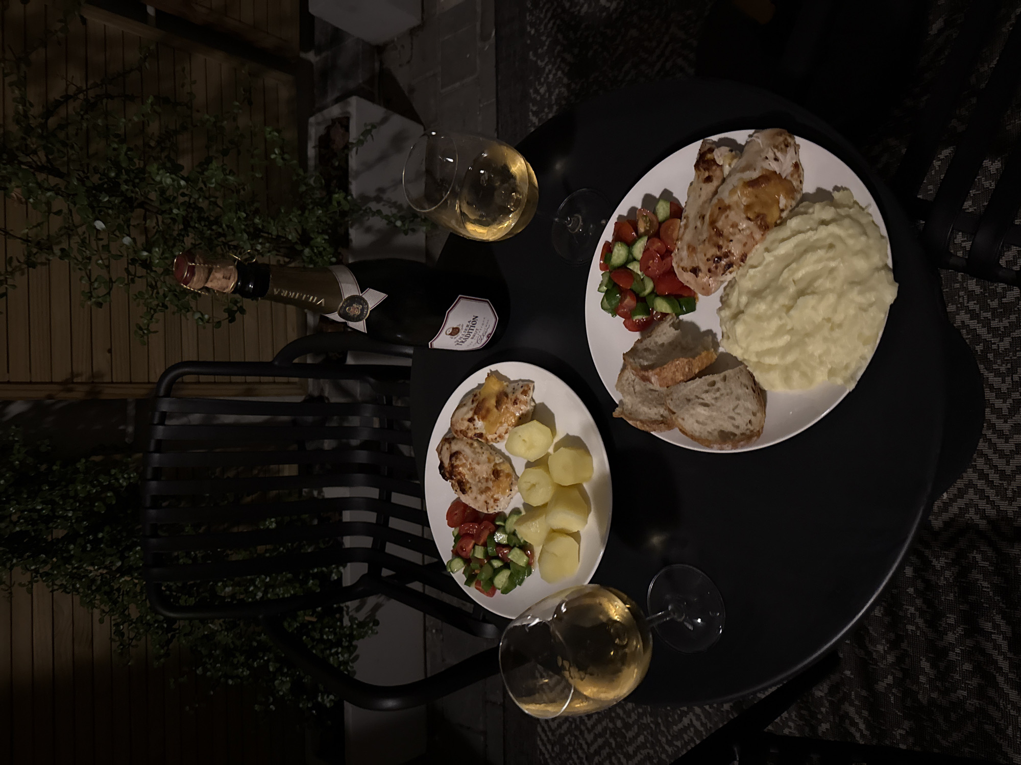 On a round, black table, outdoors, two plates of food and two glasses of white wine accompany a bottle of Villiera Tradition Brut Cap Classique sparkling wine. One plate contains cooked chicken breast, mashed potatoes, and slices of bread; the other holds cooked chicken breast, boiled potatoes, and a tomato and cucumber salad. A chair and surrounding vegetation are visible. The location is likely Cape Town, South Africa, based on the wine label.