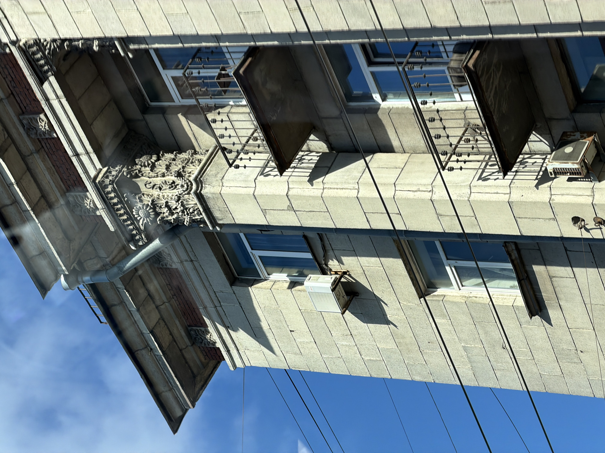 The image depicts a close-up, upward-angled view of the upper floors of a multi-story building facade under a clear blue sky. The building is constructed from large, rectangular, light-colored stone or concrete blocks, showing visible seams and some signs of age and weathering.
Architectural features include a prominent cornice at the top, detailed with red brickwork visible beneath an overhang, and decorative stone carvings. A large, ornate vertical pilaster or column on the right side of the facade is heavily embellished with carved floral motifs, specifically appearing to be sunflowers and foliage. Below the cornice, there are smaller, individual relief carvings on the stone blocks.
Multiple windows with modern white frames are visible, reflecting the blue sky. Some windows are accompanied by small, decorative wrought-iron balconies with dark railings, extending outwards from the facade. The undersides of these balcony slabs appear weathered and discolored.
A grey metal downspout runs vertically along the right side of the building, originating from the roofline. Two external air conditioning compressor units are attached to the facade: one newer, white unit on the left side, mounted on a metal bracket below a window; and an older, rusted, and discolored unit positioned further down on the right, also mounted on a bracket.
Several dark, horizontal utility lines or power cables are visible stretching across the clear blue sky against the left side of the building. The scene is brightly lit by direct sunlight, casting distinct shadows from the architectural projections, balconies, and AC units onto the building's surface, indicating the sun's position is from the upper right. No individuals are visible in the image. The specific location in Vsevolozhsk, Russia, cannot be confirmed from the visual information provided within the image.