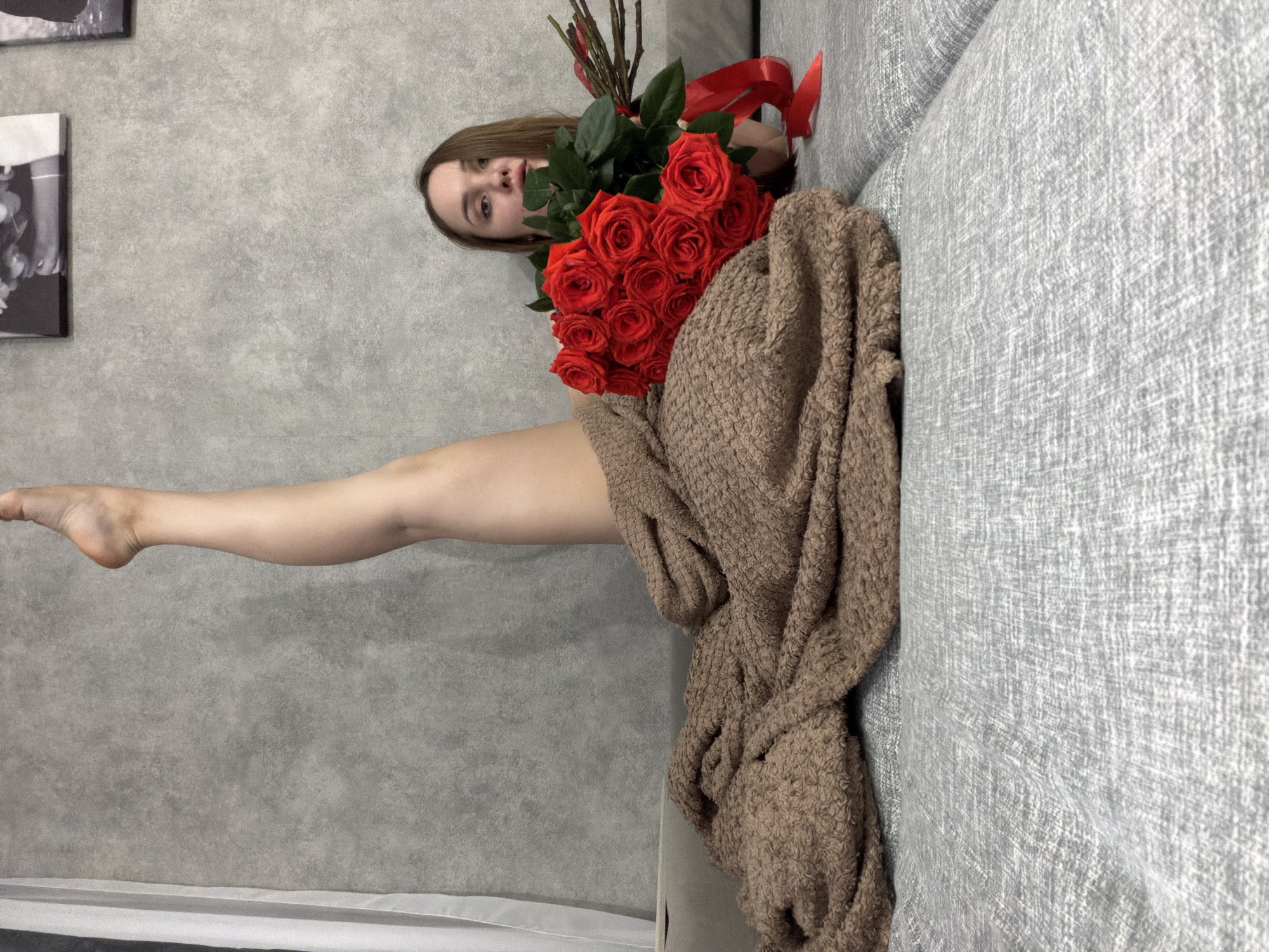 A woman is lying on a gray couch with a brown knitted blanket and a bouquet of red roses with a red ribbon. Her right leg is extended upwards towards the left. The background is a gray wall with a black and white photograph hanging on it. The location is Chaikovsky, Russia.