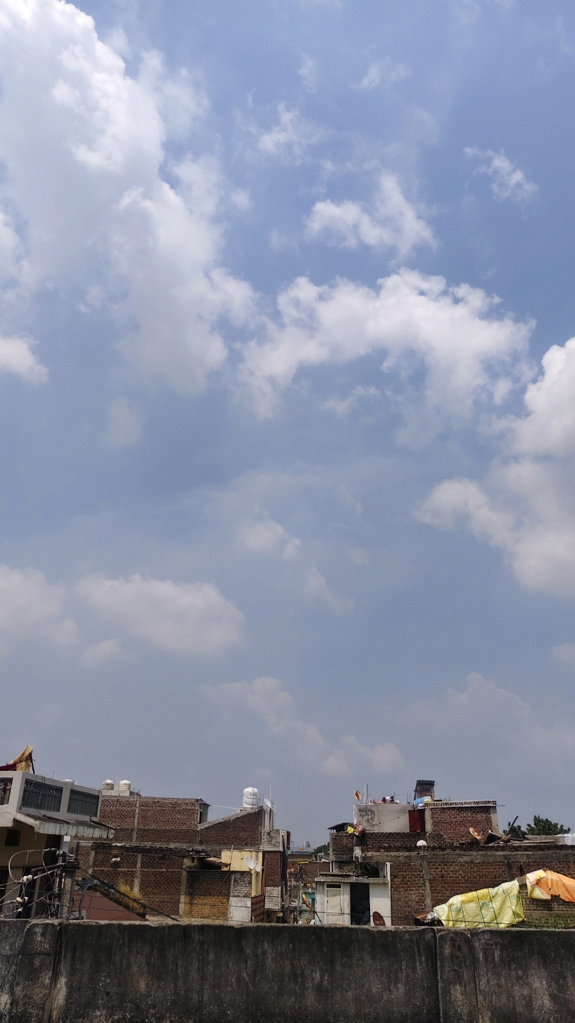 This is a vertical, eye-level, medium shot of a cityscape with a cloudy sky. The foreground is dominated by a textured, gray concrete wall. Behind the wall, a cluster of buildings, primarily made of brick and concrete, stretches across the midground. These structures vary in height and design, with some featuring exposed brickwork, satellite dishes, water tanks, and clotheslines with visible laundry. The buildings appear densely packed, characteristic of an urban environment. In the background, the sky is a pale blue with scattered white cumulus clouds, some of which are quite large and fluffy. The lighting suggests it is daytime, likely afternoon, with diffused sunlight. The image does not contain any people. The scene depicts rooftops and buildings in what appears to be a residential or mixed-use area. The location is identified as Thikri, India.