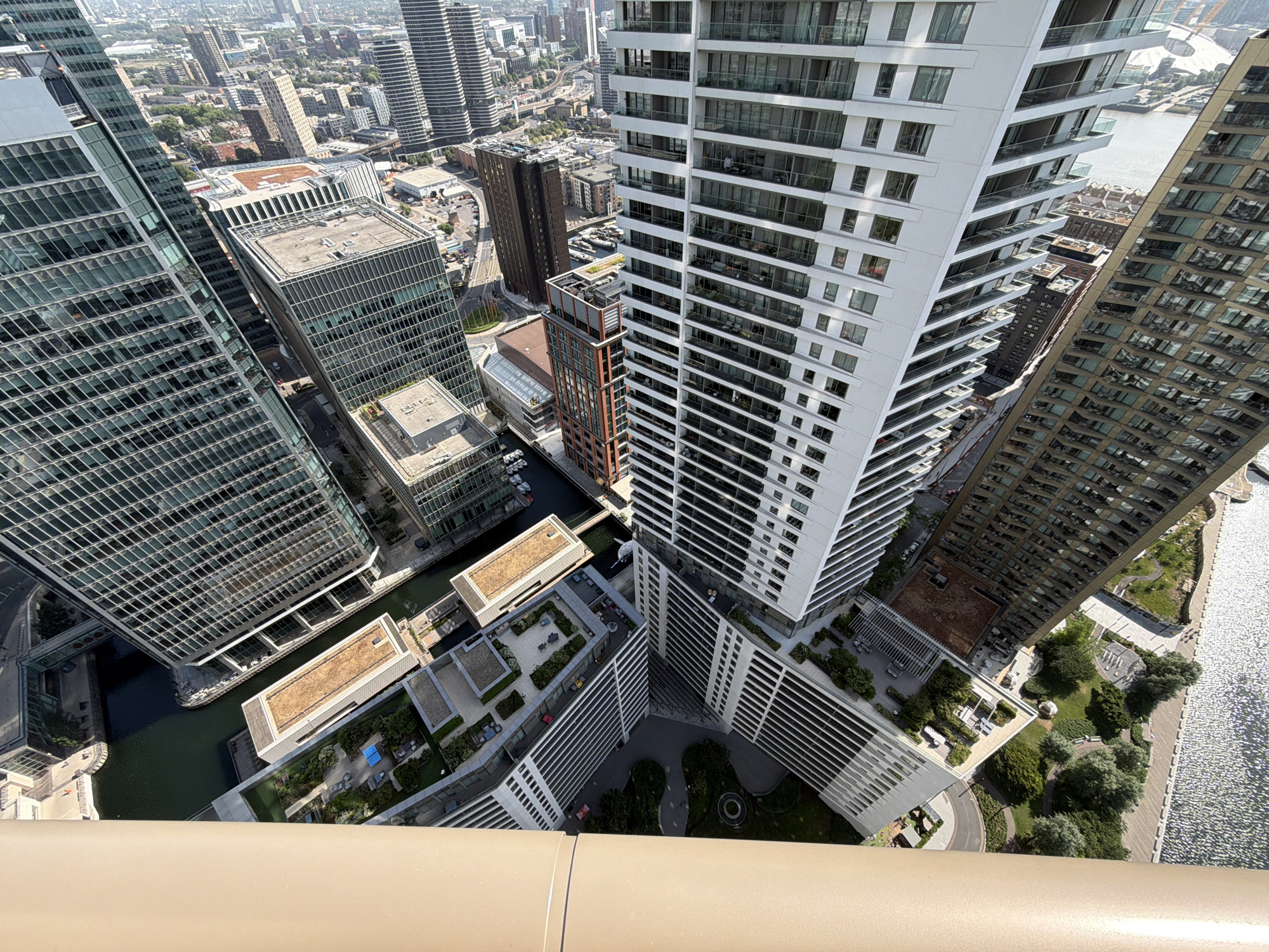 The image shows an aerial view of buildings in Canary Wharf, London, United Kingdom. The buildings are predominantly high-rise residential and commercial structures with glass and concrete exteriors. The architecture is modern with a mix of sharp angles and curved lines. The River Thames is visible to the right of the frame. The weather appears to be sunny. The viewpoint is from a high vantage point, likely another building, looking down at the surrounding cityscape.