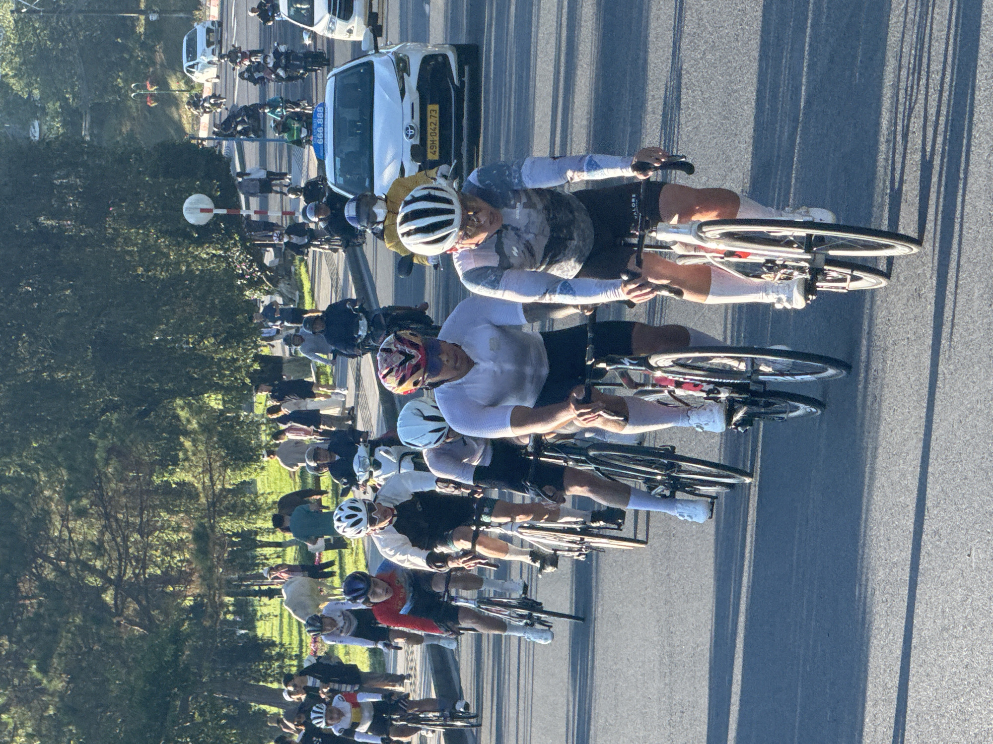 In Da Lat, Vietnam, a group of cyclists are participating in a race on a tree-lined road. The cyclists, dressed in colorful cycling gear and helmets, are riding racing bikes. One cyclist in the foreground wears a long-sleeved jersey with a grey and white abstract pattern and black shorts. To his left, another cyclist wears a white and red jersey and black shorts. Behind them, several other cyclists are visible, some on racing bikes and others on motorcycles. The road is paved, and the surrounding environment features lush green trees and vegetation. The sun is shining, casting long shadows across the road, suggesting it is either morning or late afternoon. In the background, vehicles, including cars and motorcycles, are also present on the road, indicating a public street in use. The atmosphere appears to be that of a competitive sporting event taking place in an urban or semi-urban setting within Da Lat.