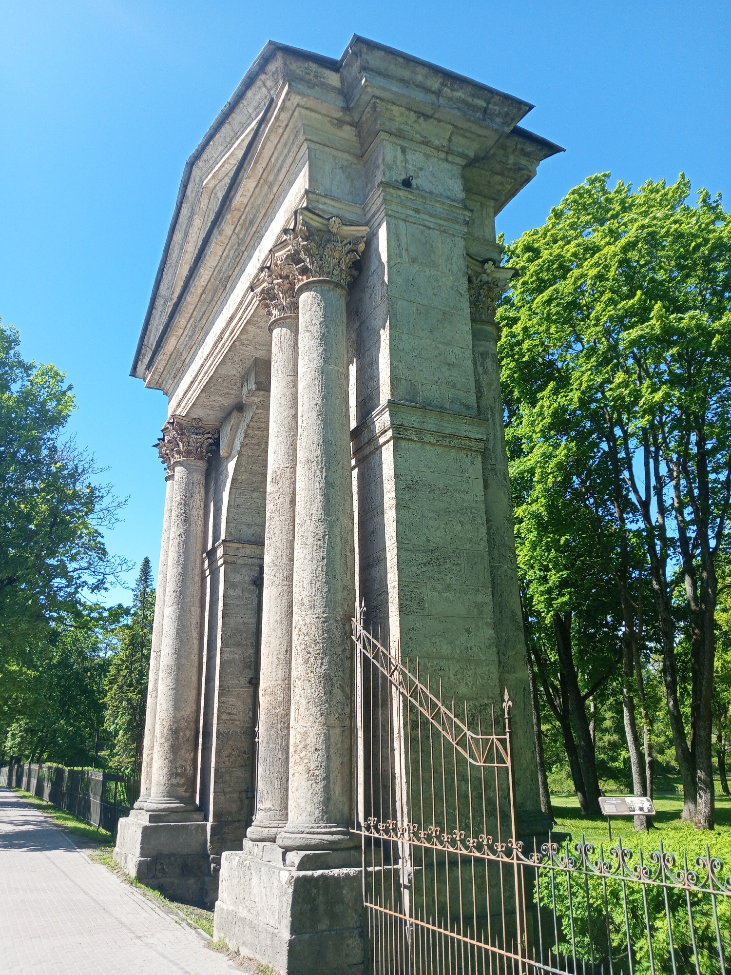 A low-angle, full shot shows the Eagle Gate in Gatchina, Russia on a bright, sunny day. The weathered, stone gate is composed of four fluted columns with ornate capitals and a flat pediment above. To the left of the gate is a paved walkway lined with a low, dark-colored metal fence with green foliage behind it. A portion of a green tree is visible in the upper left corner. To the right of the gate is a rusty, gold-colored, decorative metal fence with a grassy area and green trees behind it. A sign is visible in the grass behind the fence. The sky above is clear and blue.