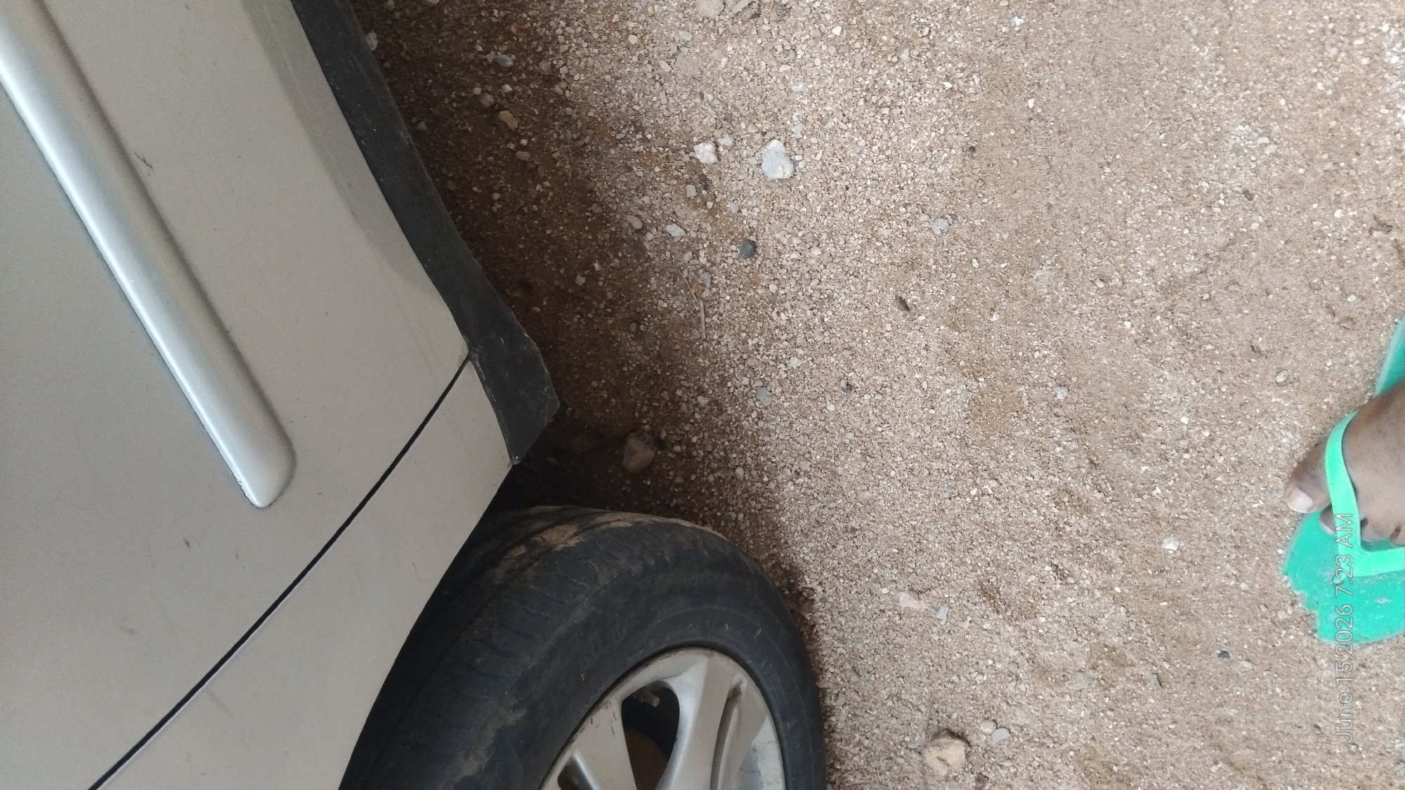 A car tire is partially visible in the upper left corner of the image, with the car's silver body and a black trim piece along the bottom. The ground is covered in small stones and dirt. In the bottom right corner, a person's foot wearing a green flip-flop is visible. The timestamp in the bottom left corner indicates the image was taken on June 15, 2026, at 7:23 AM. The scene is likely an outdoor environment, possibly a road or parking area, given the presence of a car and the ground composition. The location is identified as Howayi, Nigeria.
