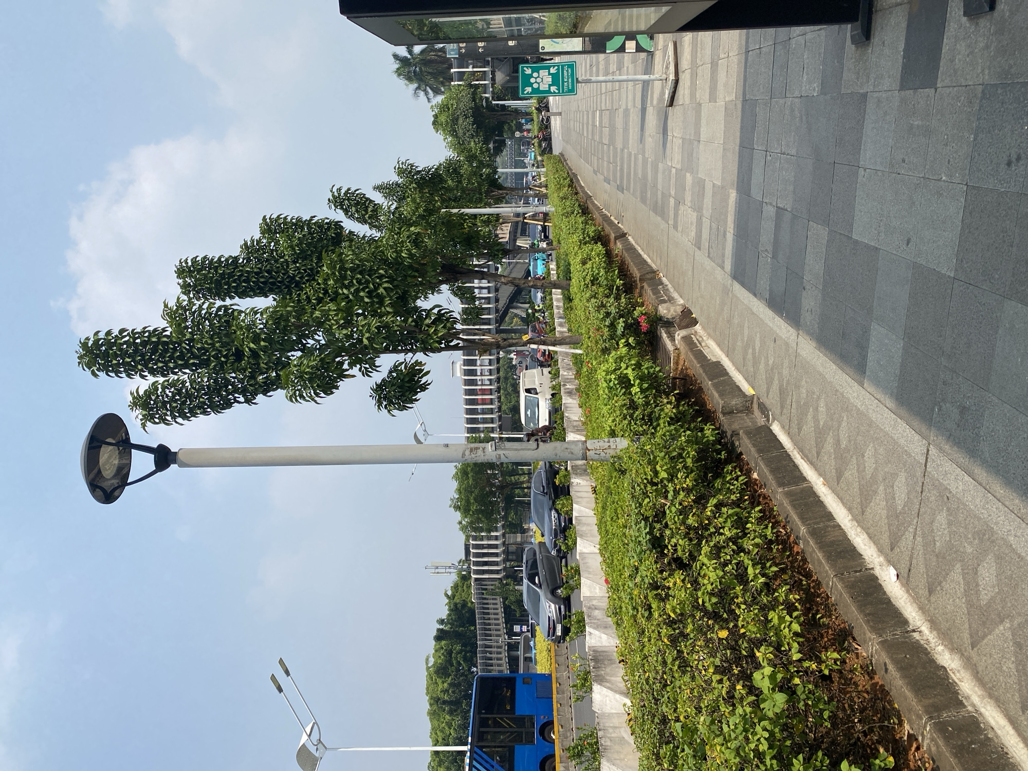 The image depicts a pedestrian walkway and adjacent road in South Jakarta, Indonesia, under a clear sky. The walkway is paved with gray square tiles and lined with manicured green shrubs and trees. Several streetlights, including a tall pole with a domed light fixture and a white pole with two directional lights, illuminate the area. On the road, a blue bus, several cars including a gray SUV and a black sedan, and a white van are visible, suggesting moderate traffic. In the background, a modern pedestrian bridge with a white and gray lattice structure spans across the road, connecting buildings on either side. A green sign with a pictogram of people and the text "TITIK KUMPUL" is positioned near the walkway, indicating an assembly point. The overall environment appears urban and well-maintained, with lush greenery contrasting with the concrete infrastructure.
