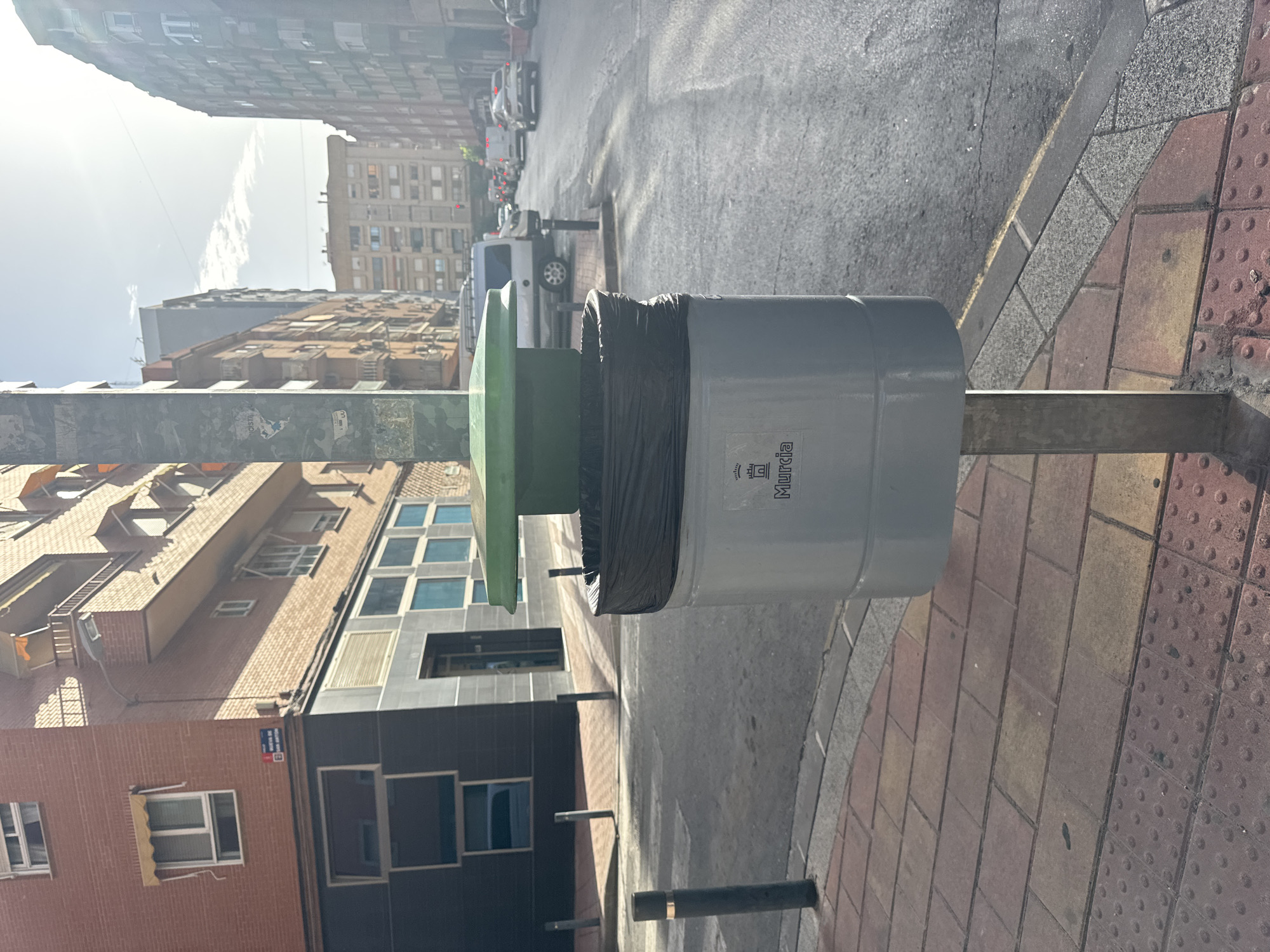 This is a photograph of a street scene in Murcia, Spain. The image is oriented vertically. The primary subject is a grey cylindrical trash can with a black trash bag protruding from the top, and a green lid. The trash can is on a metal pole. The trash can has text printed on it that appears to say "MURCIA".
The trash can is situated on a sidewalk paved with reddish-brown and yellow rectangular tiles. To the left of the trash can, there is a road with cars parked and driving. Beyond the road, there are several multi-story buildings with windows. One building in the foreground has a dark facade with large windows. Another building behind it is made of brick and has many smaller windows, some with awnings. In the background, more buildings are visible, and the sky is bright, suggesting it is daytime. The lighting indicates a sunny day.
The scene depicts an urban environment, likely a city street. There are no people visible in the frame. The overall impression is one of a typical street in a European city.