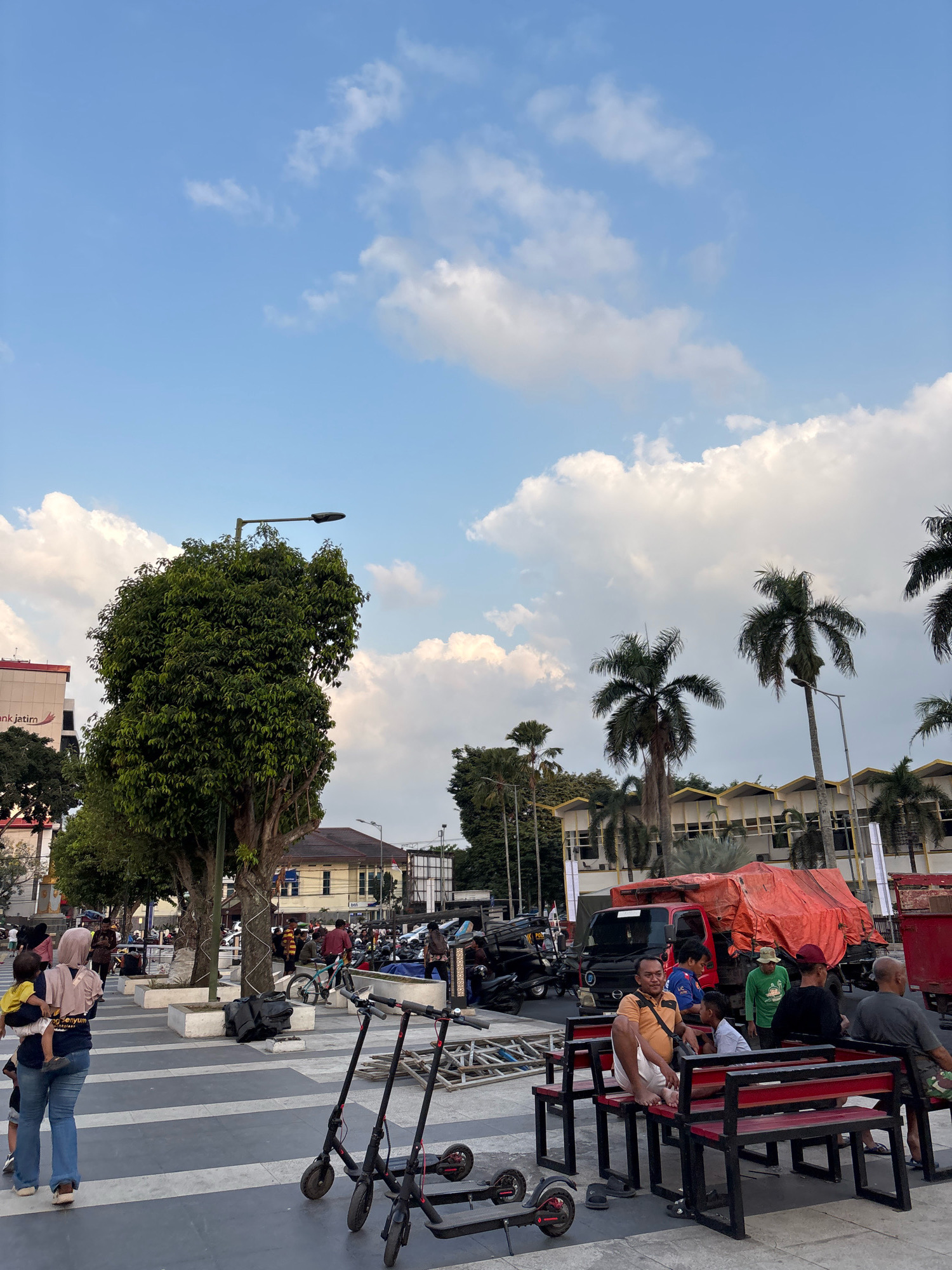 In Jember, Indonesia, a bustling public square is captured under a bright sky filled with fluffy white clouds. Several individuals are present, some walking and others seated on benches. A woman in a pink hijab carries a child on her hip, while a man in an orange shirt sits on a red bench, facing the camera. Nearby, two electric scooters are parked. In the background, palm trees sway gently, and a red truck covered with an orange tarp is visible, suggesting a busy urban environment. The architecture suggests commercial buildings with a yellow facade.