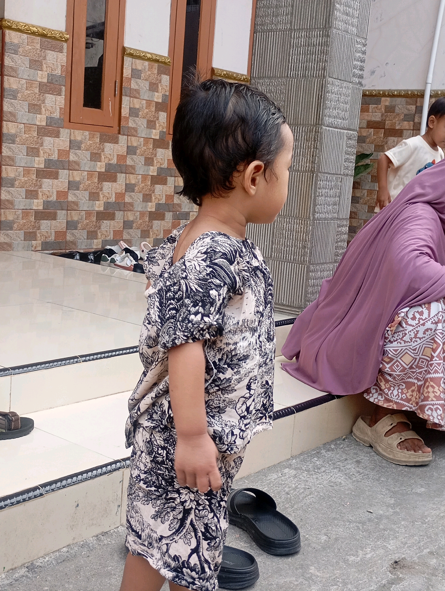 In a residential area in Babelan, Indonesia, a young child in a black and white patterned outfit stands on a concrete surface in front of a building with tiled walls and brown wooden window frames. The child is facing away from the camera and to the right. To the right of the child, a person wearing a purple hijab and a patterned skirt is seated on a step. Another child in a light-colored t-shirt is visible in the background. Several pairs of shoes are scattered on the ground near the building's entrance, and a sandal is placed on the step. The overall environment appears to be an outdoor walkway or patio area of a house.