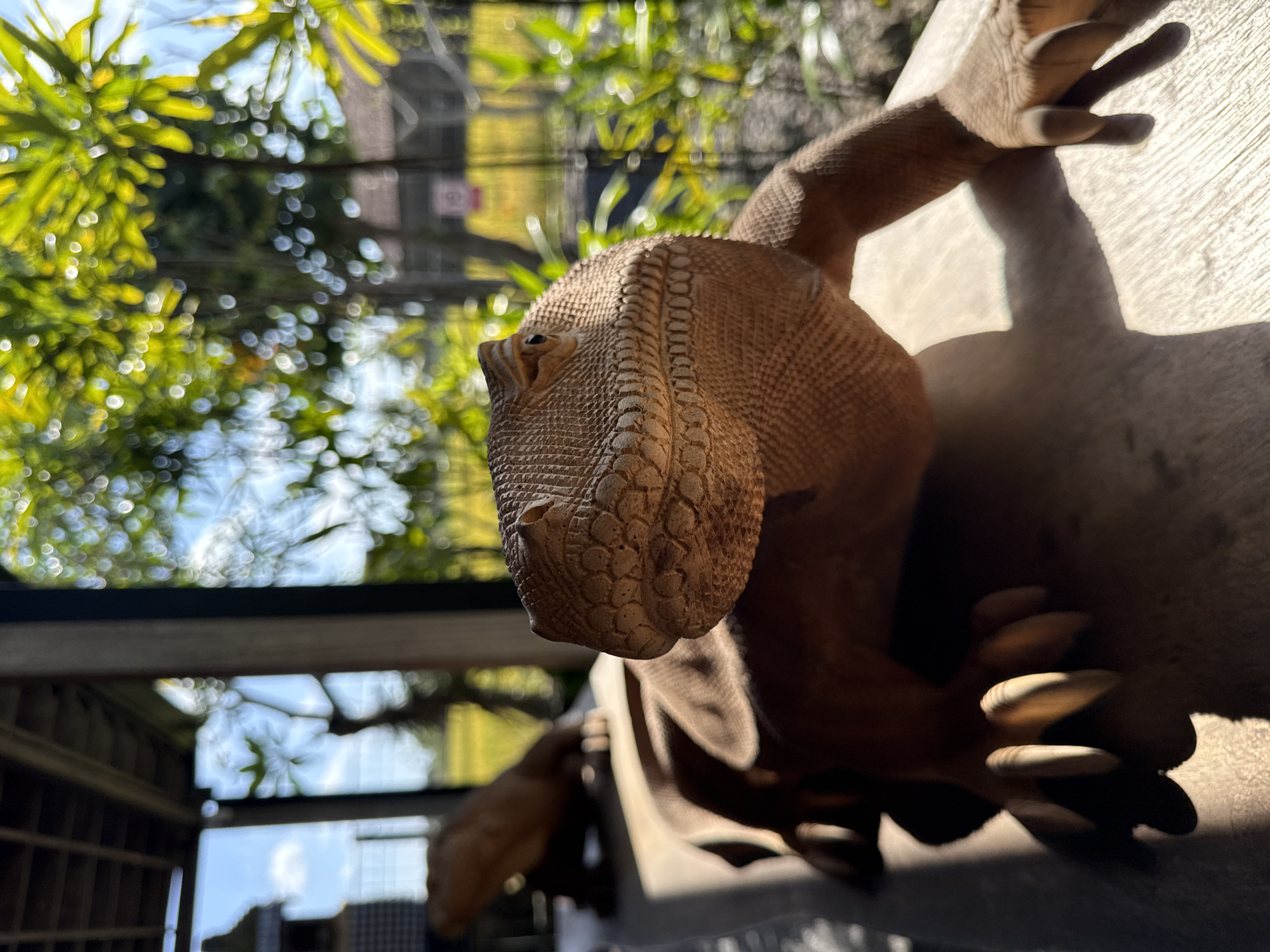 A wooden sculpture of a lizard is positioned on a wooden surface in the foreground, with another similar sculpture blurred in the background. The lizard sculpture in the foreground is facing left, with its head tilted slightly upwards. The lighting suggests it is outdoors during the day, with dappled sunlight falling on the sculpture and creating shadows. In the background, out of focus, are lush green foliage, a wooden structure that appears to be a roof or pergola, and a glimpse of a bright yellow field, possibly a rice paddy. The scene evokes a serene, natural environment. Given the context, this scene is likely located in a craft village or artisan shop in Sukawati, Indonesia, known for its wood carvings and traditional crafts.