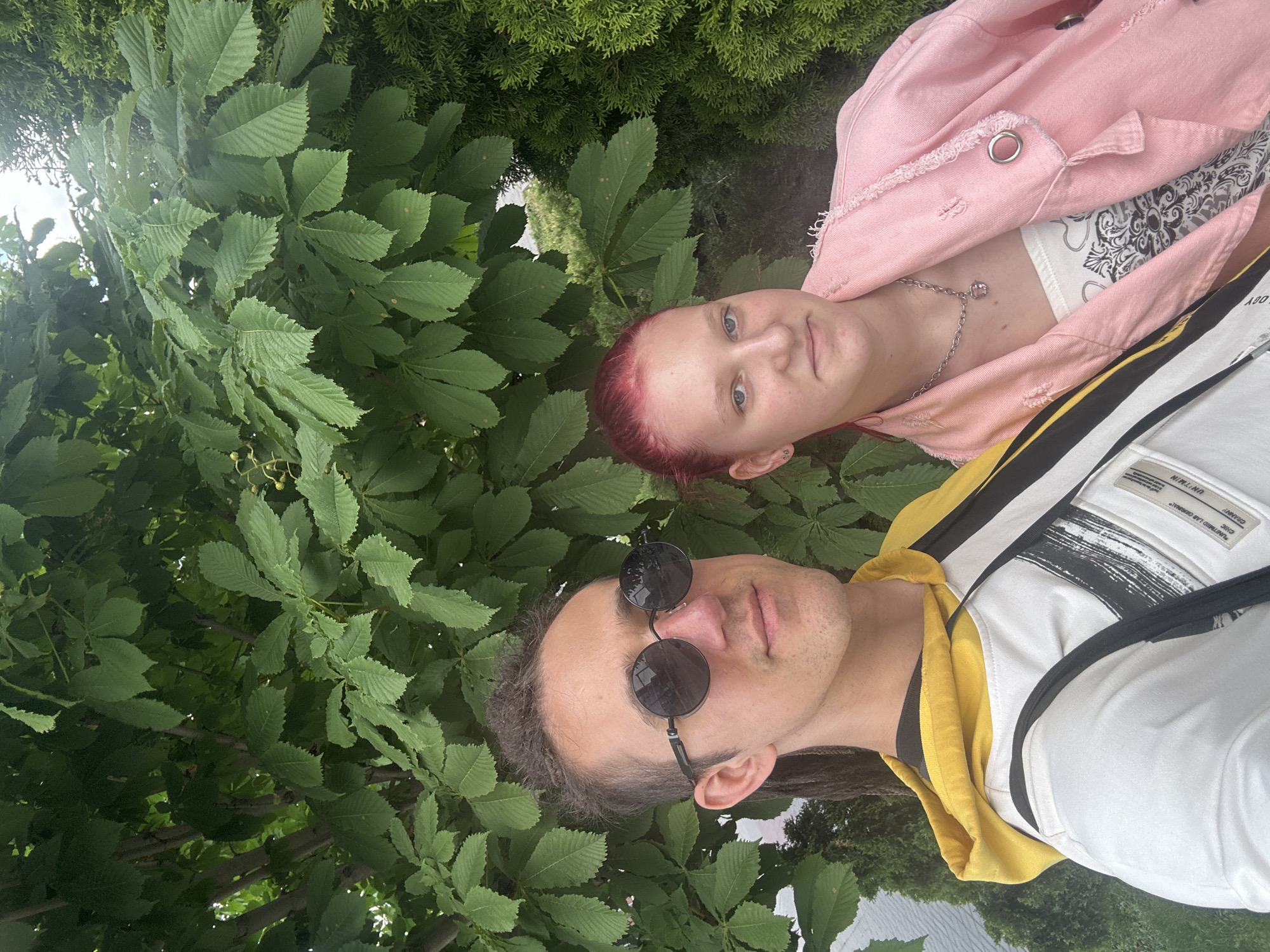 A selfie shows two individuals against a background of dense green foliage, likely trees. A person with short red hair, wearing a pink denim jacket over a white patterned top and a silver chain necklace, is positioned in the upper right of the frame. Below and to the left is another person wearing round sunglasses, a yellow hooded shirt, and a white shirt with black stripes, with a black strap across the shirt, possibly from a bag.
The location cannot be confirmed as Saratov, Russia.