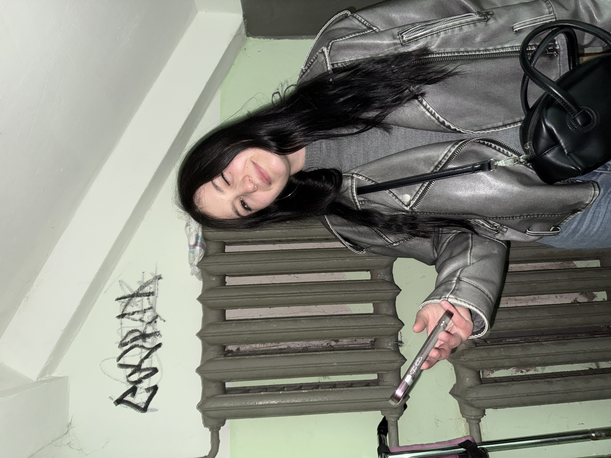 A young woman with long, dark hair, wearing a grey jacket and jeans, stands in what appears to be a stairwell or entryway, potentially in Chaikovsky, Russia, though the city's identification is not directly confirmed within the image. She holds a smartphone in her right hand. A cast-iron radiator is visible behind her, along with graffiti on the wall to the left. A black purse hangs from her shoulder. The environment appears utilitarian and possibly public.