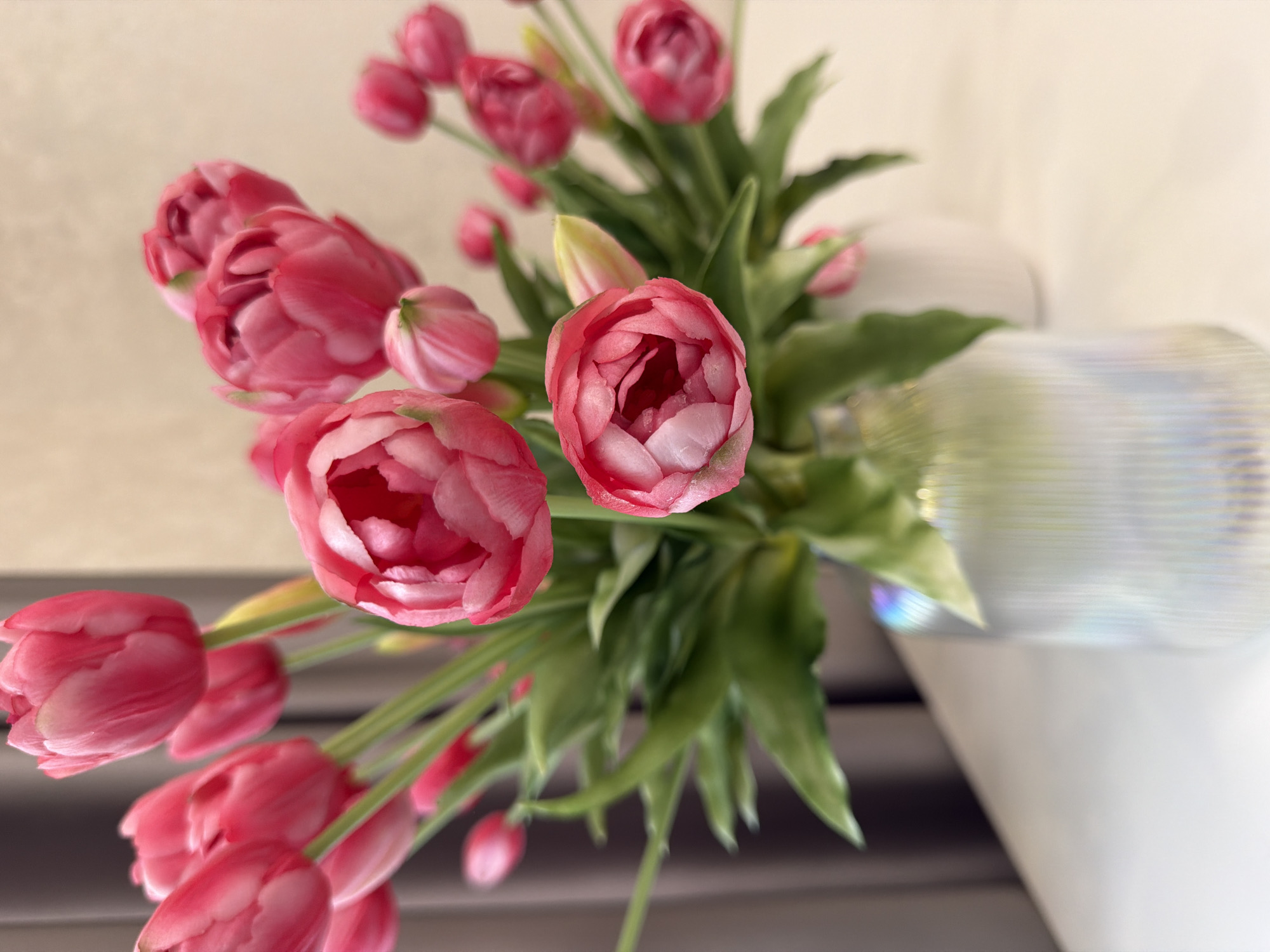 A bouquet of pink tulips is displayed in a vase on a white surface. The tulips are a mix of fully bloomed and budding flowers, with vibrant green leaves. The vase appears to be made of textured glass with an iridescent finish. In the background, there is a blurry curtain and a wall. The overall scene suggests an indoor setting, possibly a home or a professional space, with soft lighting. The image does not provide any specific details about a city or location within a city.