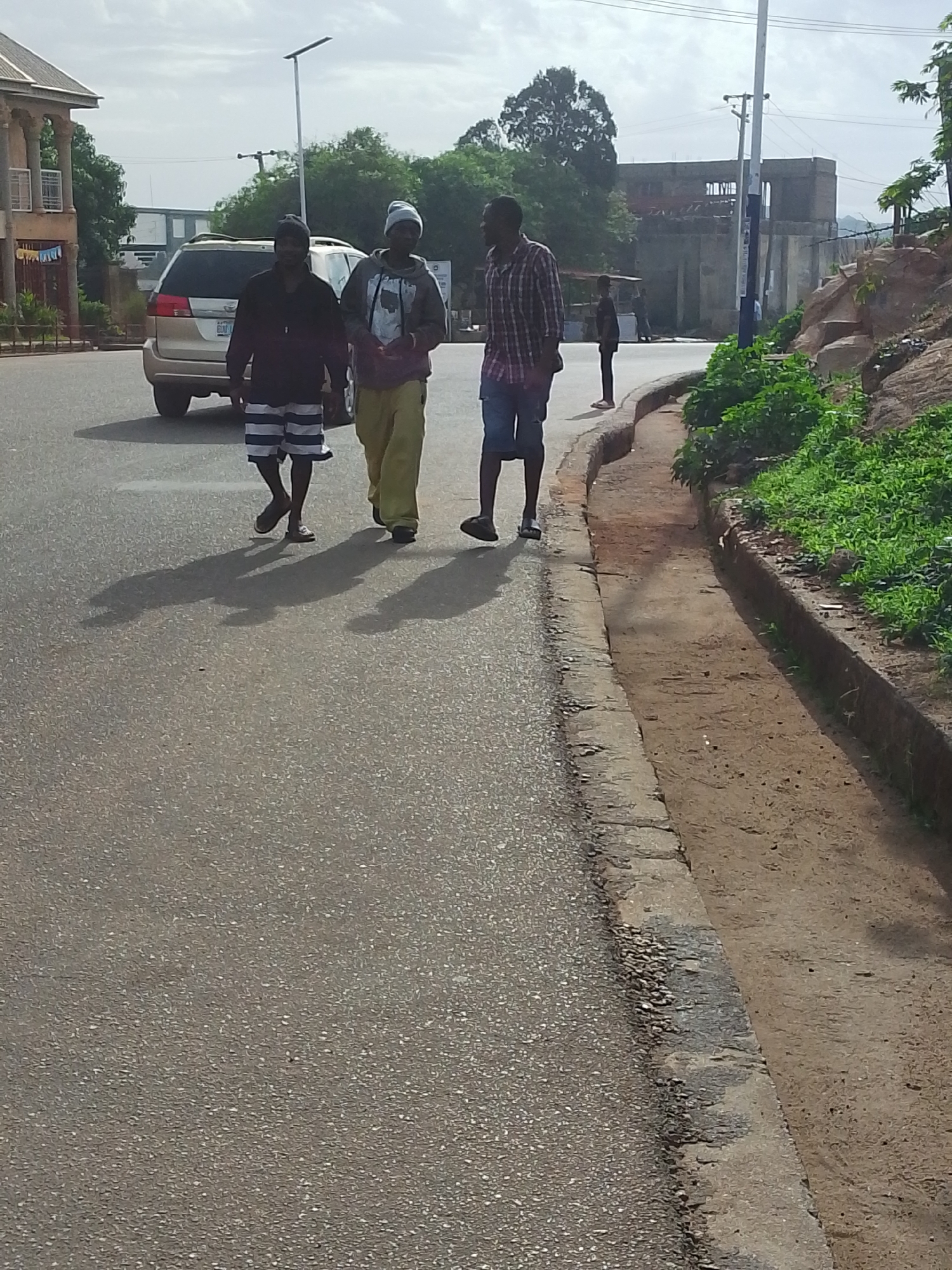 Three men are walking on a paved road in Jos, Nigeria. The man on the left wears a dark long-sleeved shirt and striped shorts. The man in the center wears a grey hooded sweatshirt, yellow pants, and a beanie. The man on the right wears a red plaid shirt and blue shorts. A tan SUV is partially visible in the background to the left of the group. A fourth person stands further down the road. A concrete drainage ditch borders the road on the right, adjacent to a rocky area with green vegetation. Buildings and trees are visible in the background.