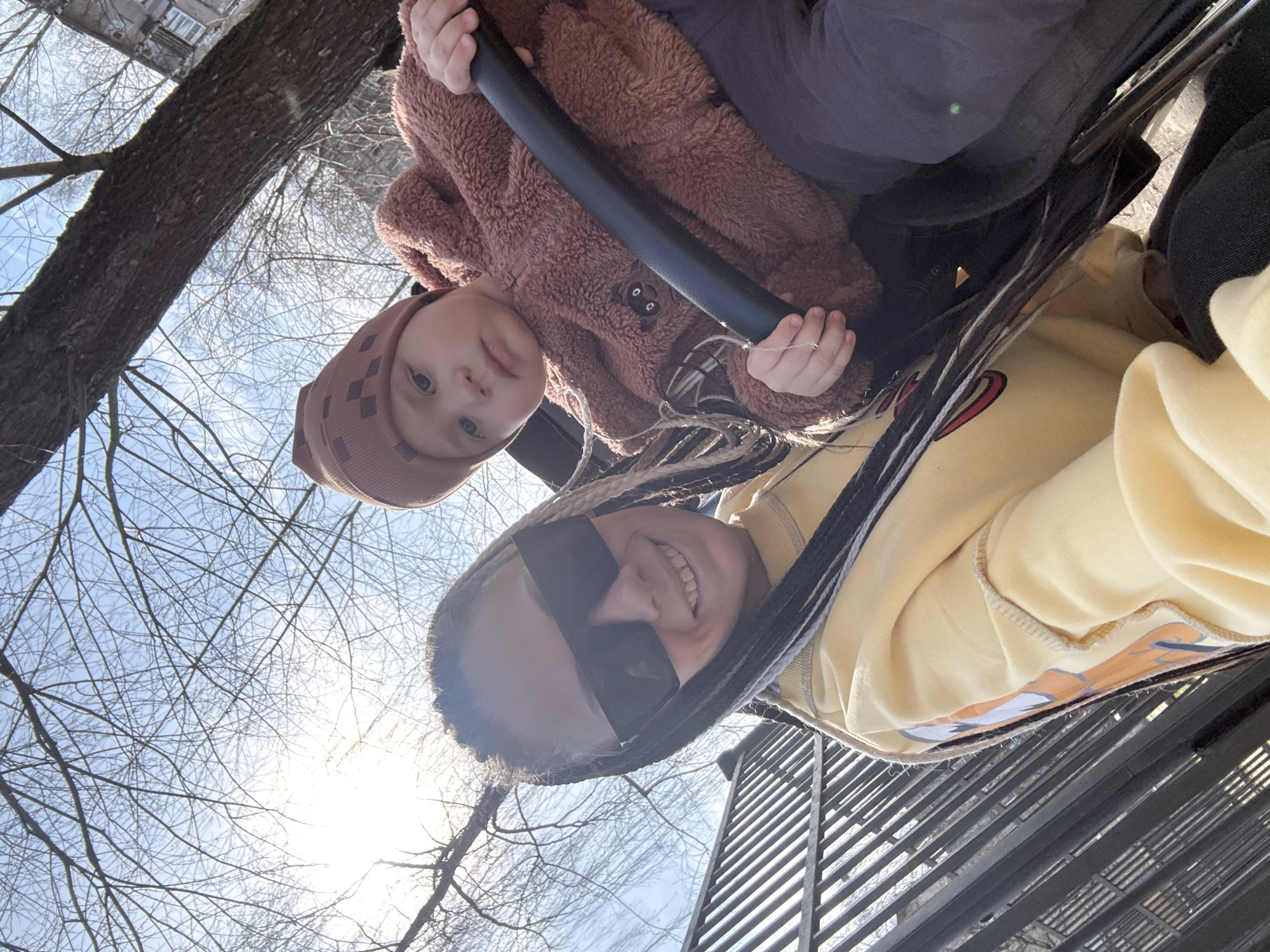 A low-angle shot shows a child and an adult together in an outdoor setting. The child, wearing a brown hat and a textured brown jacket, is seated and holding onto a black bar. The adult, wearing a yellow sweatshirt and sunglasses, is also seated and smiling. Both appear to be on some kind of ride or swing.
The background includes bare tree branches against a pale sky with visible sunlight. Elements of a building are partially visible, suggesting an urban area. A metal structure with repeating parallel bars is also present in the foreground. The image is rotated counter-clockwise. The exact location in the city of Rusanovo, Russia is indeterminable from this image.