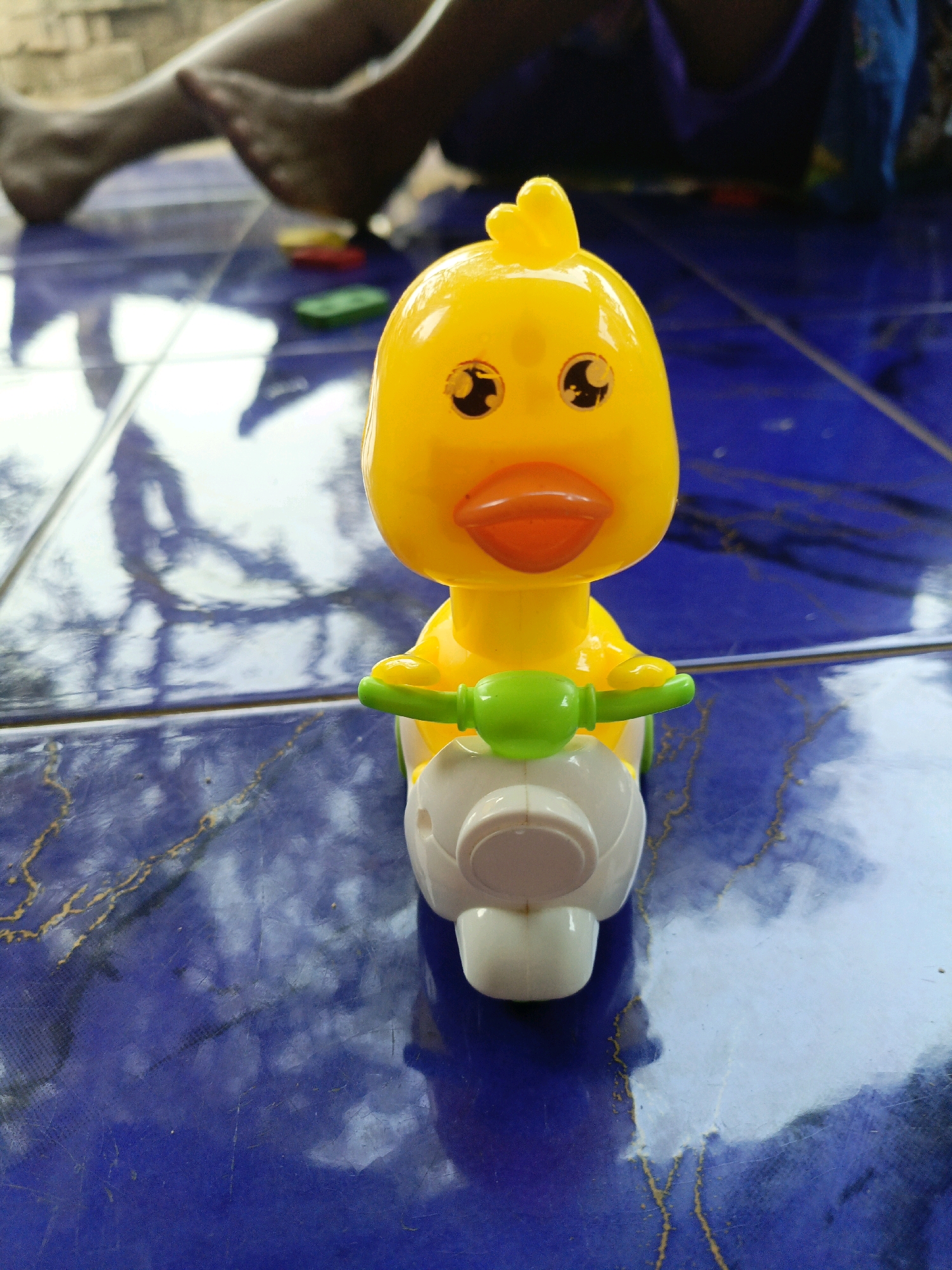 A yellow plastic duck toy rides a white toy motorcycle. The duck has black eyes with a sparkle effect, an orange beak, and a small yellow crest on its head. The motorcycle has green handlebars and a white body. The toy is positioned on a blue tiled surface that has a reflective sheen and a marbled pattern. In the background, blurred legs and feet of a person are visible, along with some small colorful objects on the floor, including a yellow, red, and green item. The lighting suggests an indoor or shaded outdoor environment. The scene appears to be located in Jenu, Indonesia, likely in a residential or outdoor play area.