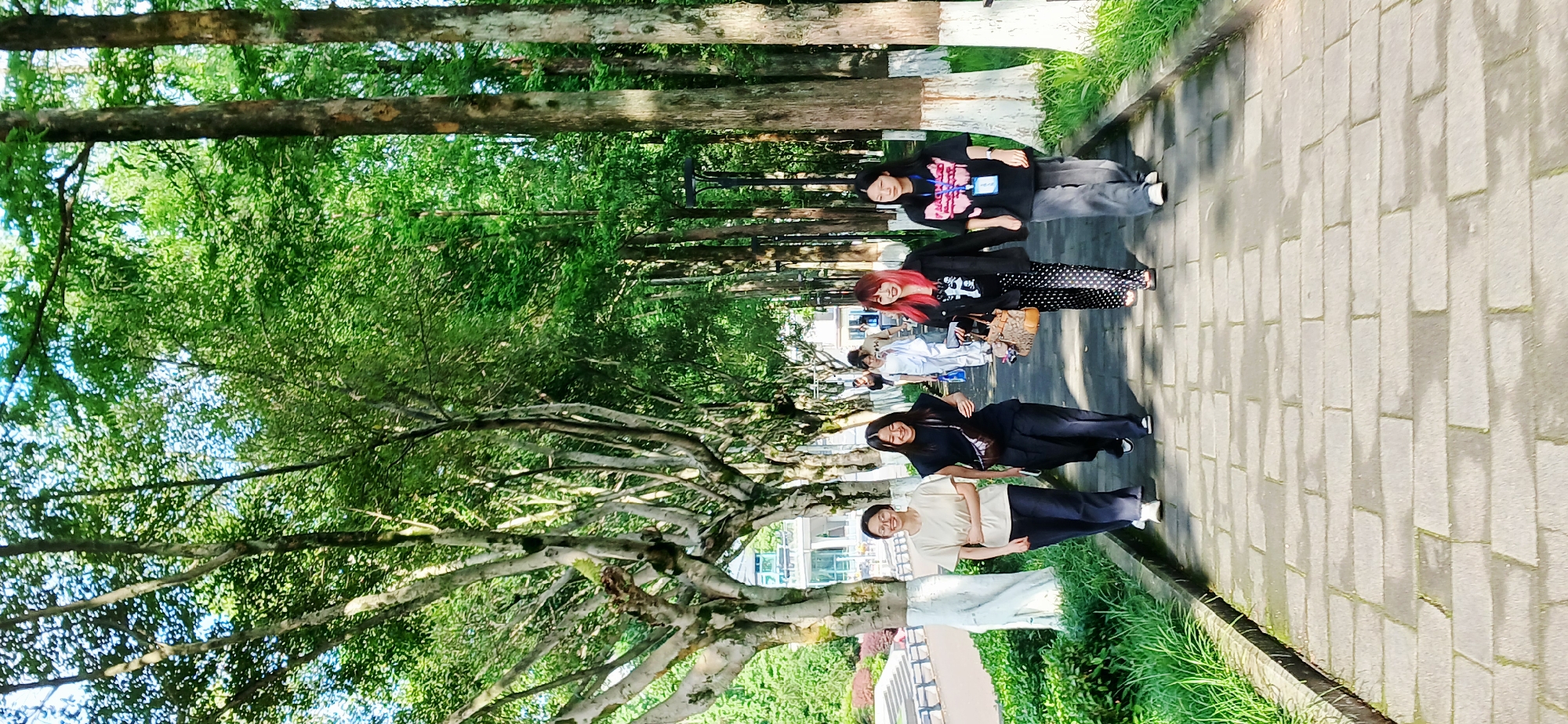 The image shows a group of five people standing on a brick-paved sidewalk under a canopy of trees. The location is likely in the Chongqing High-tech Zone, China, given the environment and information.
The person closest to the viewer is wearing a light-colored shirt and dark pants. The next person is wearing black clothes. The third person has red hair, a black jacket, and polka dot pants, carrying a brown bag. The fourth person has a black top. The fifth person is wearing a black sweatshirt with a graphic design and grey pants.
The sidewalk runs parallel to a row of trees with green foliage overhead, suggesting a park or a tree-lined street. Sunlight is filtering through the trees, creating shadows on the sidewalk. A curb separates the sidewalk from a grassy area with other trees and shrubs. In the background, a building can be seen.