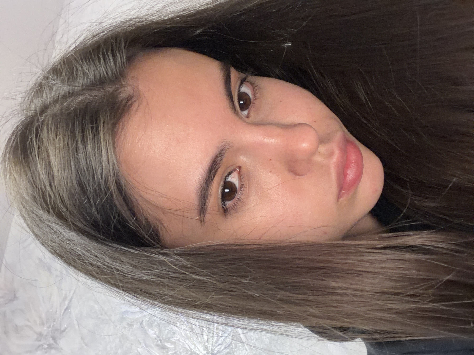 A close-up, slightly rotated image depicts a young woman with long, dark brown hair that has ashy blonde highlights near the roots. Her skin is fair, with a neutral undertone and minimal visible blemishes. Her eyebrows are dark and well-defined. Her eyes are brown. Her nose is average in size and shape, and her lips are full and pink.
No information suggests the location is Chaikovsky, Russia. The background is blurred and indistinct, but appears to be a textured white surface.