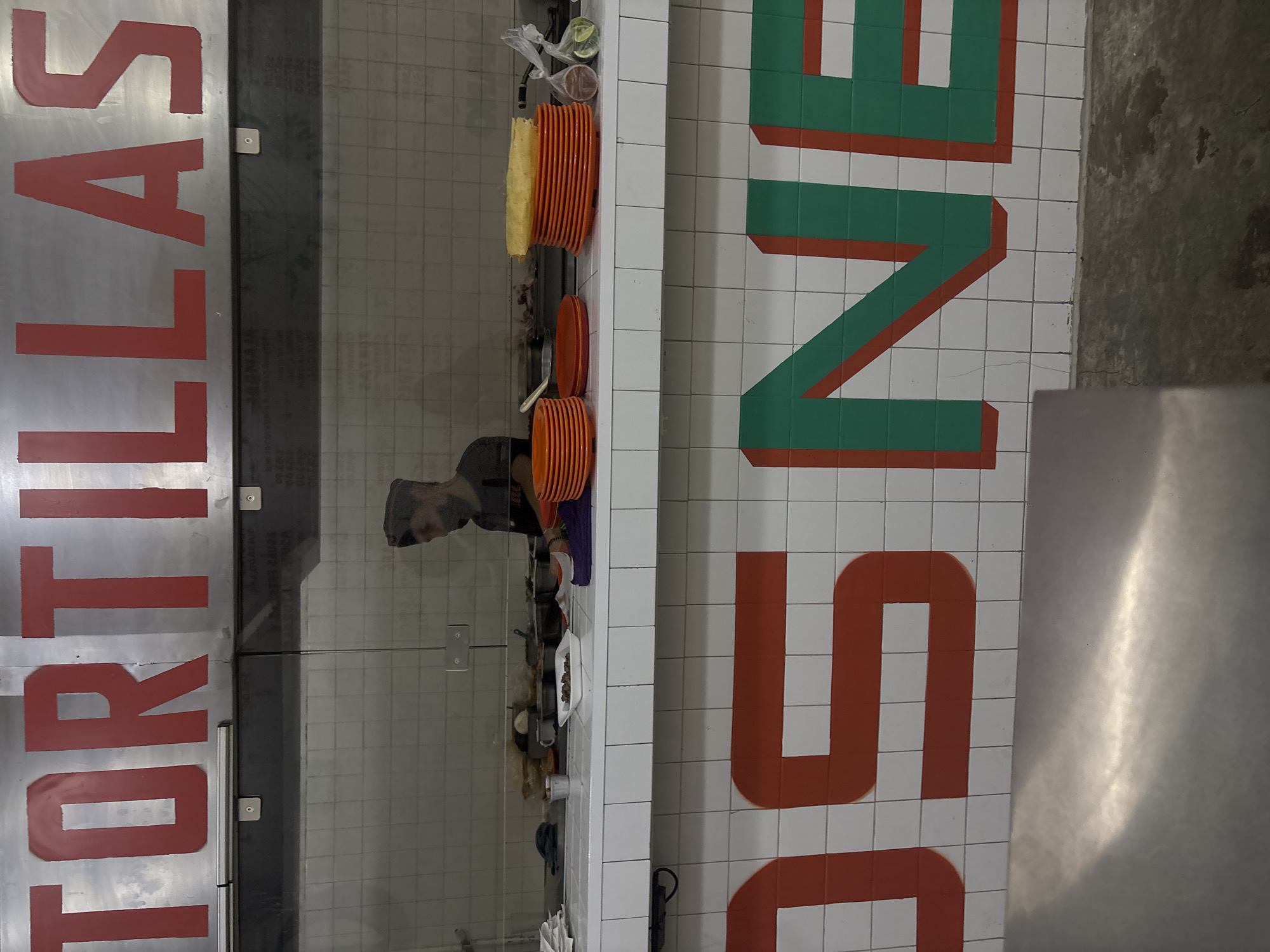 The image captures an indoor scene, likely a food preparation or serving area in a restaurant or market. On the left side, a large sign with a silver background displays the word "TORTILLAS" in bold red capital letters, running horizontally across the top of the frame. Below this sign, a clear glass partition separates the viewer from a counter.
Through the reflection in this glass partition, a person is visible behind the counter. This individual, possibly a chef or food worker, has short dark hair and a beard, wearing a dark cap and a dark top. They appear to be engaged in an activity behind the counter, though the specifics are obscured by the reflection.
The counter itself, which is white, holds several items. Stacks of bright orange plates of various sizes are neatly arranged, ready for use. A yellow sponge or cleaning pad is also visible, along with other kitchen containers and utensils.
To the right of the counter, a tiled surface, possibly a floor or lower wall, features a distinctive graphic design. This design incorporates large, blocky letters in green and red against a white tiled background. Part of the word "SINE" is discernible in red, with green lines forming portions of the letters, suggesting another sign or branding element. The tiles create a grid pattern across this surface. On the far right, a shiny, metallic surface is partially visible. The setting suggests a clean and organized culinary space, possibly in Mexico City given the context of "TORTILLAS."