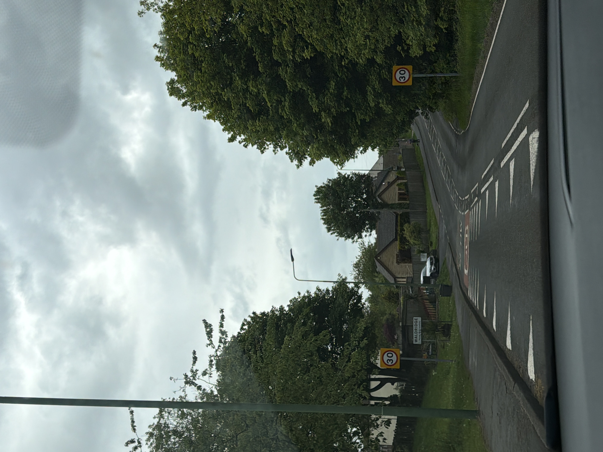 A street scene in Waldridge, a village in the city of Durham, United Kingdom, is captured from the perspective of a vehicle. The overcast sky suggests it could be a typical day in the region. The road is marked with white dashed lines towards the center and solid white lines with adjacent chevrons along the curb, indicating lane markings. Two prominent yellow circular road signs reading "30" signify a 30 miles per hour speed limit. Lush green trees line the roadside. Buildings, including houses, are partially visible behind the vegetation. A street sign reading "WALDRIDGE" is also visible, confirming the location. A silver car is parked along the road. A lamp post stands along the road.