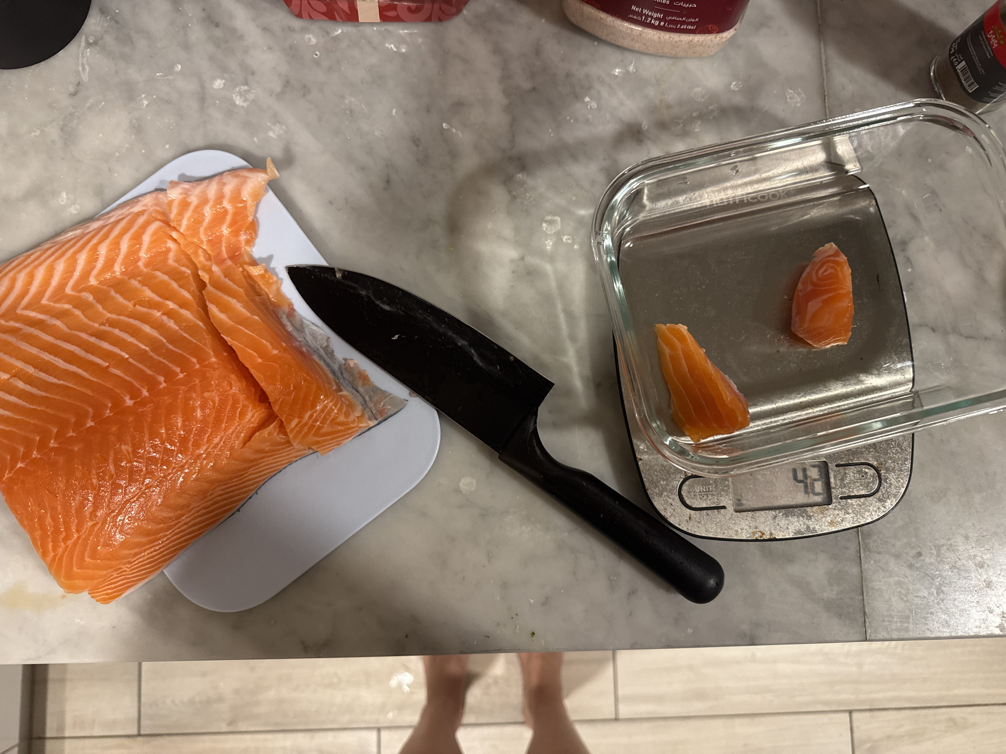 A still life photograph shows a slab of raw salmon on a light blue cutting board to the left, a black handled knife lying diagonally across the scene, and a clear glass dish on a digital scale reading "42" to the right. The salmon slab appears to be cut into pieces, with two pieces already separated and placed in the glass dish. Beside the dish are pink salt and a jar of dried pepper. The objects are placed on a marble-like countertop. The lower part of the image reveals a person's feet. The location is not specified, but text on the salt jar mentions Arabic words suggesting the United Arab Emirates.