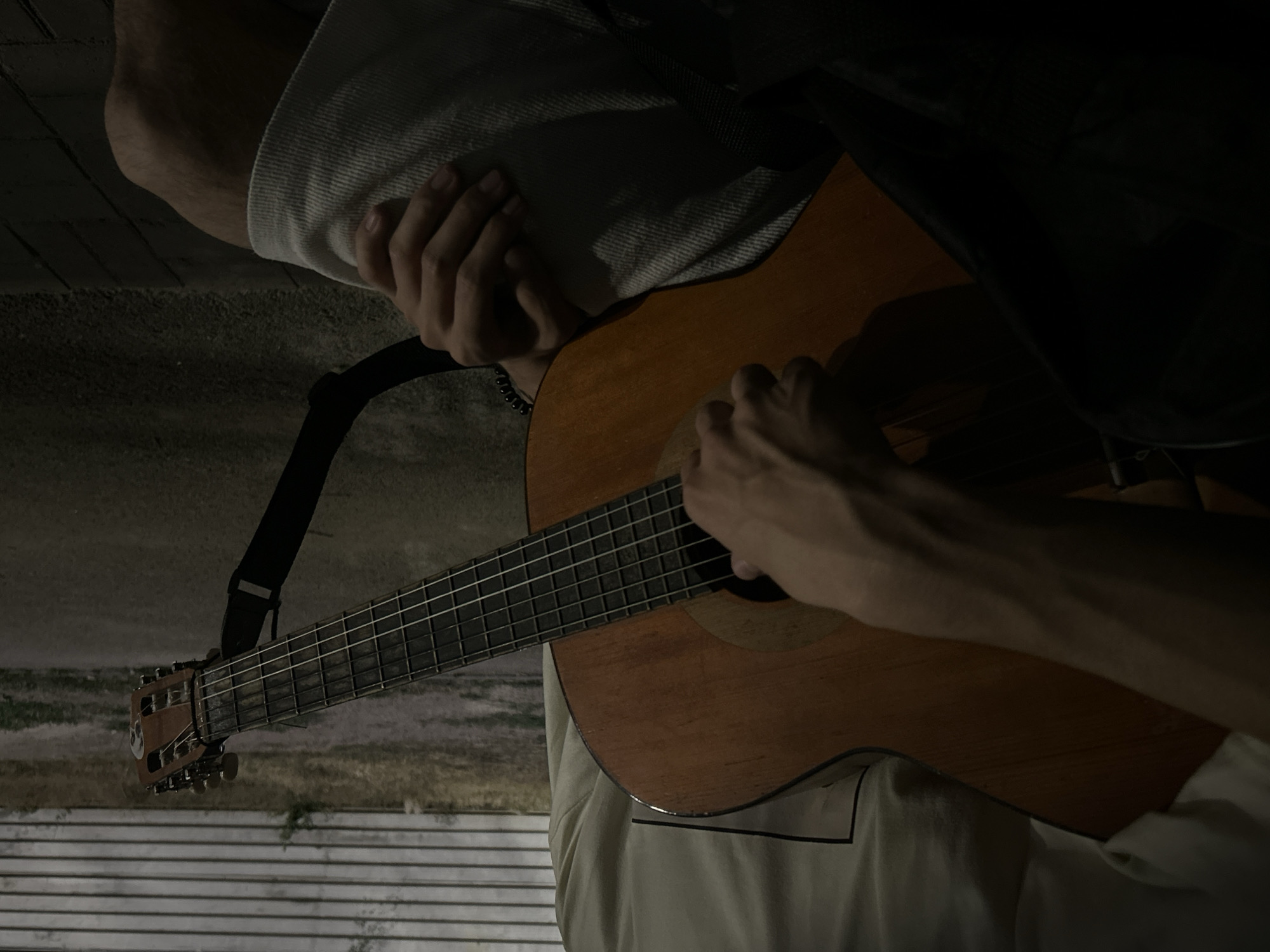 A close-up shot shows a person playing an acoustic guitar. The guitarist's hands are visible on the fretboard and the strings. The guitar has a wooden body and a black strap. The person is wearing a light-colored t-shirt and light-colored shorts. The background is out of focus but appears to be an outdoor setting with a fence or wall and some vegetation. The lighting suggests it is dusk or nighttime. The scene is located in Taraz city administration, Kazakhstan.