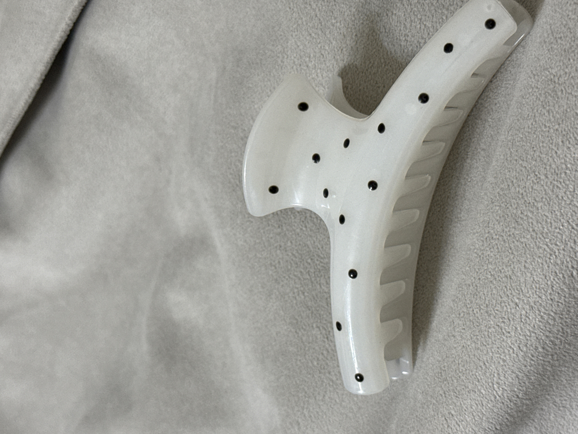 A white, opaque hair claw clip, featuring a glossy finish and numerous small, irregularly placed black polka dots, is positioned diagonally in the frame. Its design includes an upper, broader section with a subtle curve and a lower, claw-like part with visible internal teeth, indicating it is slightly open or designed to display its gripping structure.
The hair clip rests on a soft, light gray textile surface, likely a blanket or fleece fabric, which constitutes the entire background. The fabric displays a consistent, gentle texture and subtle folds, particularly noticeable on the left side of the image, contributing to slight variations in tone. The lighting is soft and even, illuminating the object and its textured backdrop without creating harsh shadows.
The scene is static, offering a close-up view of the hair accessory in what appears to be an indoor setting. There are no people, specific activities, or discernible text present in the image. The provided location context is Naberezhnye Chelny, Russia.