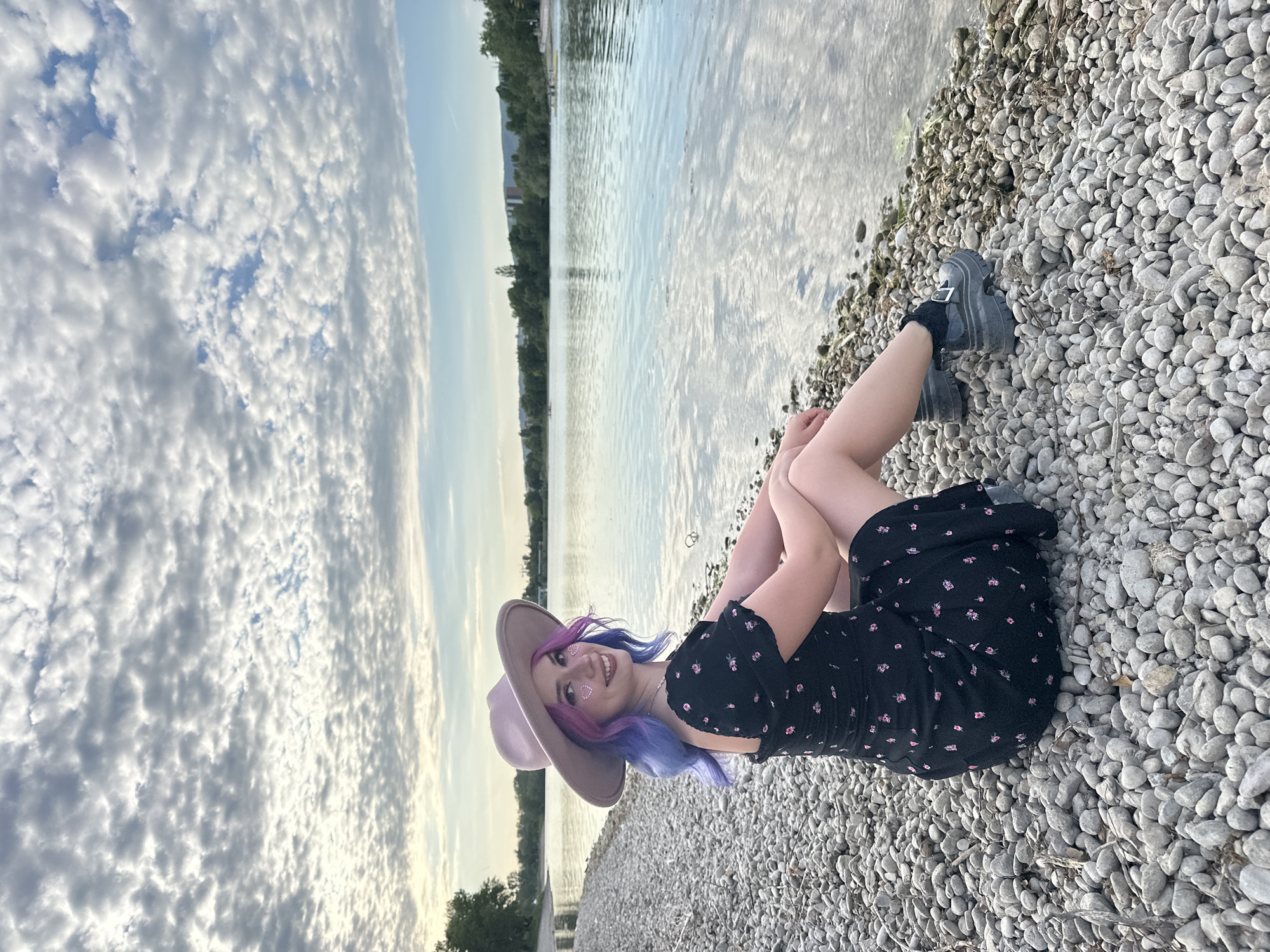 The image, displayed in a portrait orientation but originally captured in landscape and rotated 90 degrees clockwise, presents a young woman posing cheerfully on a pebbly shore by a body of water, likely in the City of Zagreb, Croatia. She is lying back with her legs bent, resting on the smooth grey and white pebbles that form the shoreline.
The woman features striking, two-toned hair, transitioning from purple to pink. She wears a light brown, wide-brimmed fedora-style hat. Her attire includes a dark-colored, short-sleeved dress adorned with small, light-colored floral patterns, and chunky dark boots with buckles, paired with black socks. Her face is turned towards the camera, revealing a wide, joyful smile, and she has small, heart-shaped embellishments on her cheeks.
Behind her, a calm body of water stretches into the distance, reflecting the muted tones of the sky. The far bank is lined with dense green trees and hints of distant buildings, suggesting an urban fringe near nature. The sky above is filled with a distinctive pattern of textured white clouds against patches of pale blue, indicating a partly cloudy late afternoon or early evening. The overall scene conveys a relaxed and pleasant atmosphere. No visible text is present.