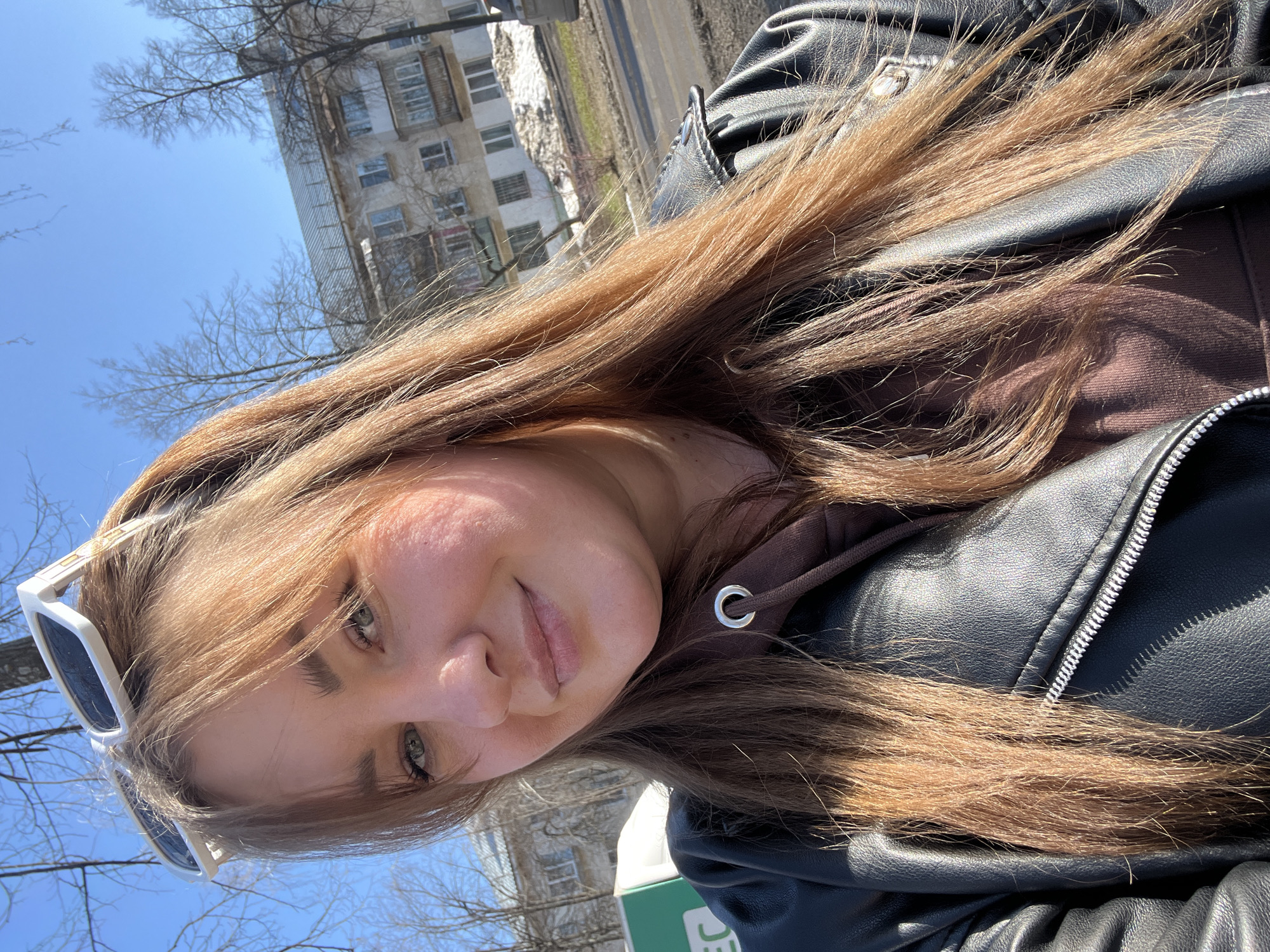 A selfie depicts a person with long, light brown hair and fair skin, wearing a dark brown hooded sweatshirt beneath a black leather jacket. White-framed sunglasses are positioned on their head. The person is smiling subtly at the camera.
In the blurred background, a multi-story building typical of Soviet-era architecture is visible. The presence of leafless trees suggests either late autumn or early spring. Considering the architectural style and the style of the bus stop to the left, the location is likely in the city of Chaikovsky, Perm Krai, Russia. The subject is likely standing near a bus stop, given the visible green signage. A road is partially visible to the right side of the background. The sky is clear and blue.