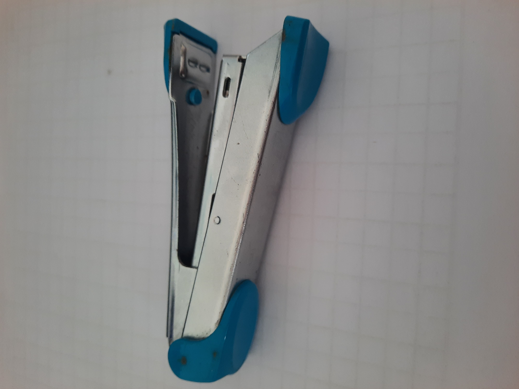 A silver and blue stapler is lying on a white grid paper. The stapler is positioned horizontally, with its front end pointing to the left and its rear end to the right. The stapler has a silver body with blue plastic accents at both ends. The top arm of the stapler is lifted, revealing the staple mechanism inside. The grid paper appears to be a notebook or a planner, with faint gray lines forming squares. The background is a plain white surface. There are no people or other objects visible in the image. The scene is well-lit and the focus is on the stapler.