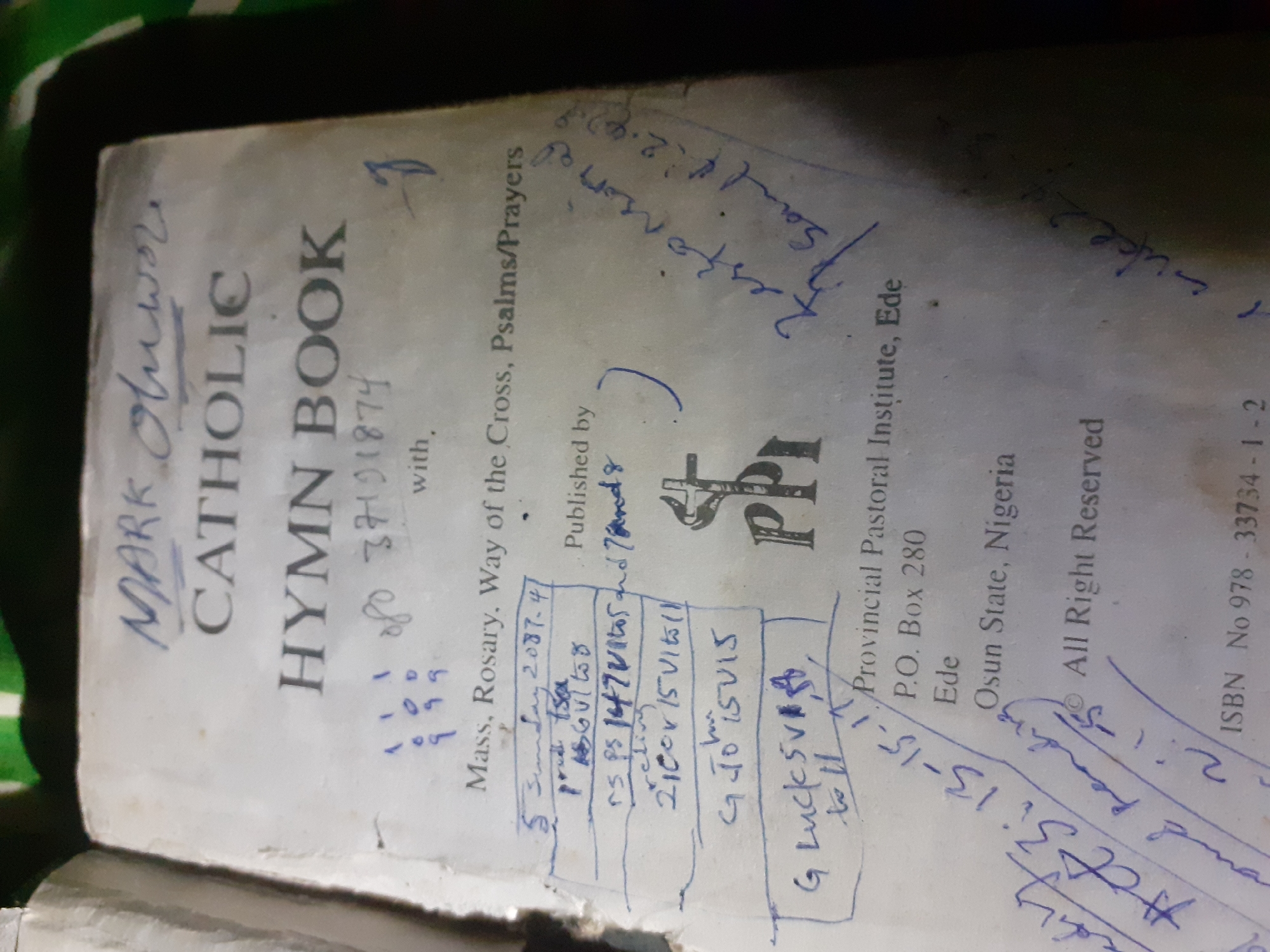 A close-up, slightly angled, overhead shot shows the cover of a worn, white "CATHOLIC HYMN BOOK" with blue handwritten text. The title of the book, "MARK Olwor / CATHOLIC / HYMN BOOK", is at the top. Below this, handwritten in blue ink, are the numbers "1 1 1 / 0 0 D / 9 9 9", followed by "of 37191874", and then "with." The body of the book cover has various handwritten notes and markings in blue ink, including lists of numbers, dates, and what appears to be a list of chapters or sections, like "5 Sunday 2087-4", "ps 147Vl05 and 7mnd 8", "relin", "2100r 15Vl05(1)", "G Jo 15Vl5", and "G Luck 5V 88, to 11". There is also a drawing of what looks like a chalice with a cross on top. To the right of the book title, a large, looping, blue-ink swirl or signature is present. Further down on the right side, more blue ink writing is visible, with words like "response", "Psalm 1:2-2-9", and "luke 24". On the bottom right, the text "Provincial Pastoral Institute, Ede / P.O. Box 280 / Ede / Osun State, Nigeria" is printed. Below this, "All Right Reserved" is also printed. The bottom edge of the book shows an ISBN number: "ISBN No 978 - 33734 - 1 - 2". The background is dark and out of focus, with some hints of green. The image does not provide any information about the city of Jalingo, Nigeria.